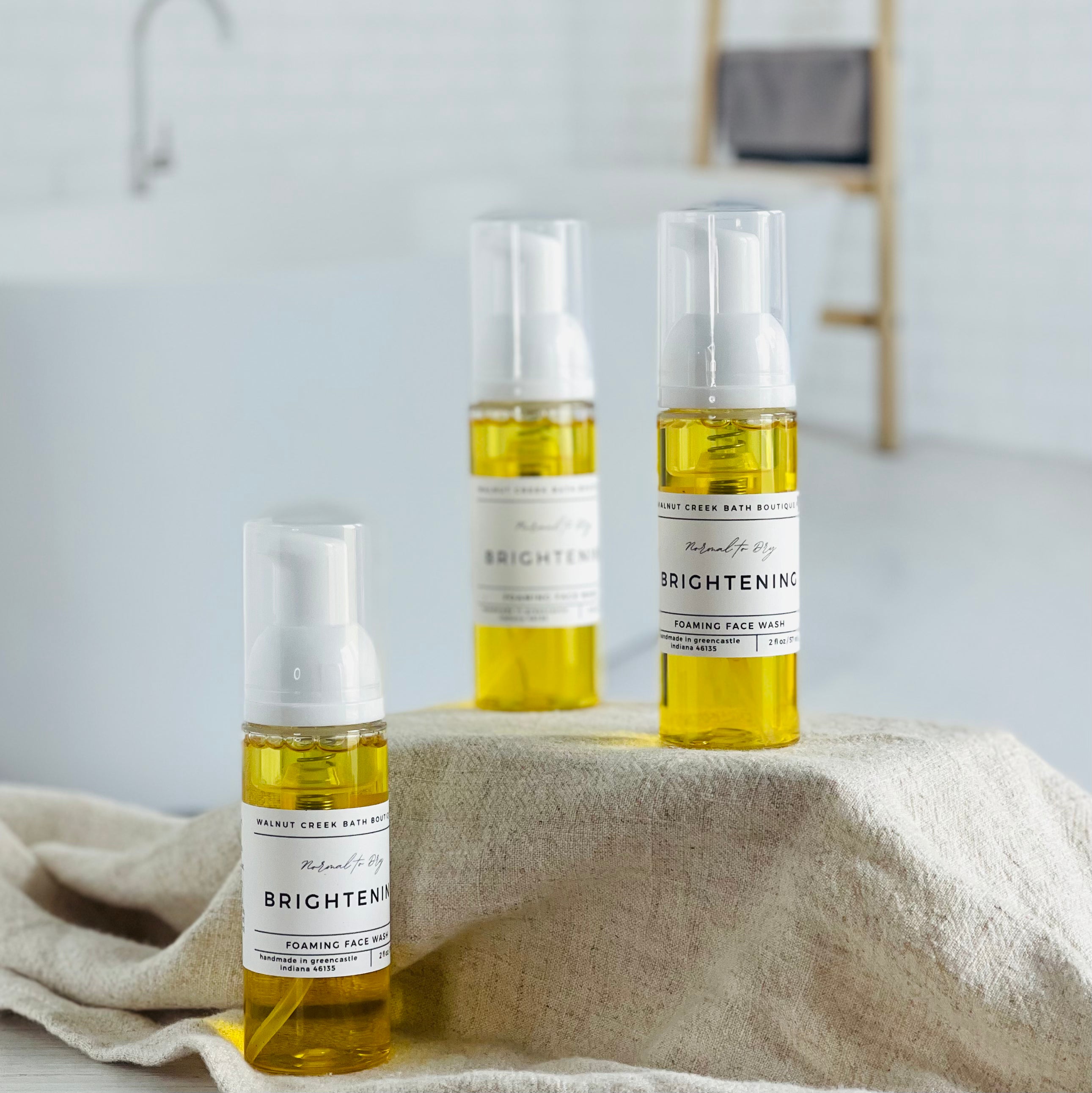 3 brightening foaming face washes are shown, 2 are on a tan cloth covered riser and then there is one on the base closest to you. they are a lovely bright yellow color. in the background is a spalike bathroom and a sage green vase with florals