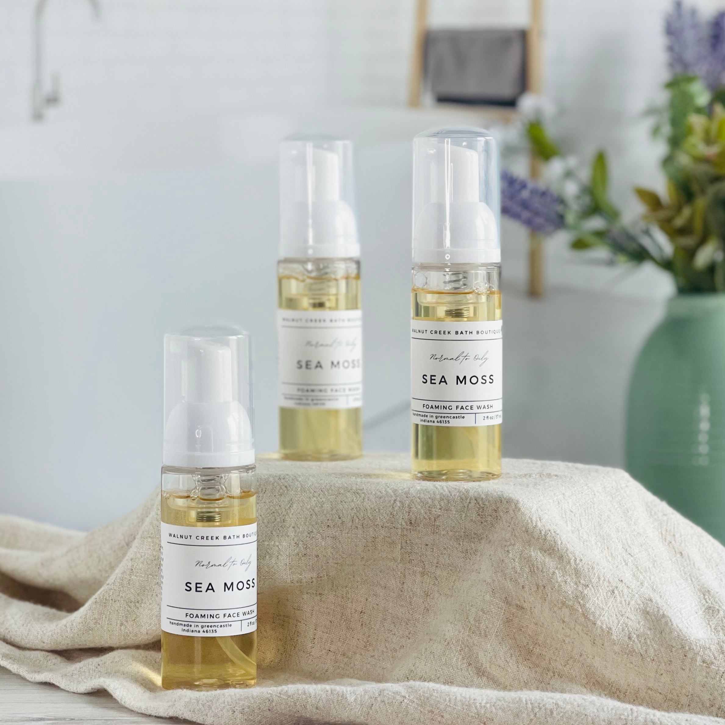 3 sea moss foaming face washes are shown, 2 are on a tan cloth covered riser and then there is one on the base closest to you. they are a light yellow color. in the background is a spa like bathroom and a sage green vase with florals