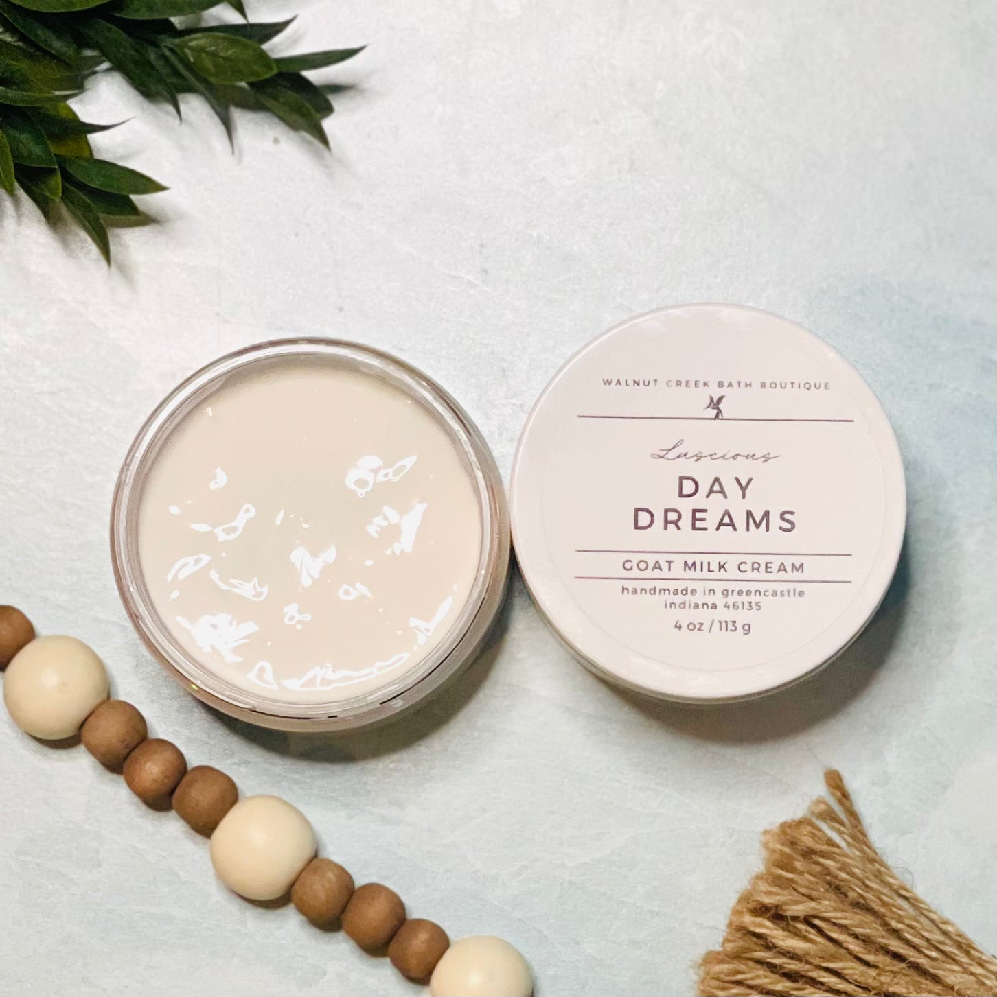 4 oz day dreams goat milk cream open with the lid next to it. the texture is so creamy and luscious looking. there is some greenery on the top left and some wooden beads running along the edge with the tassel peaking just in view on the bottom right.