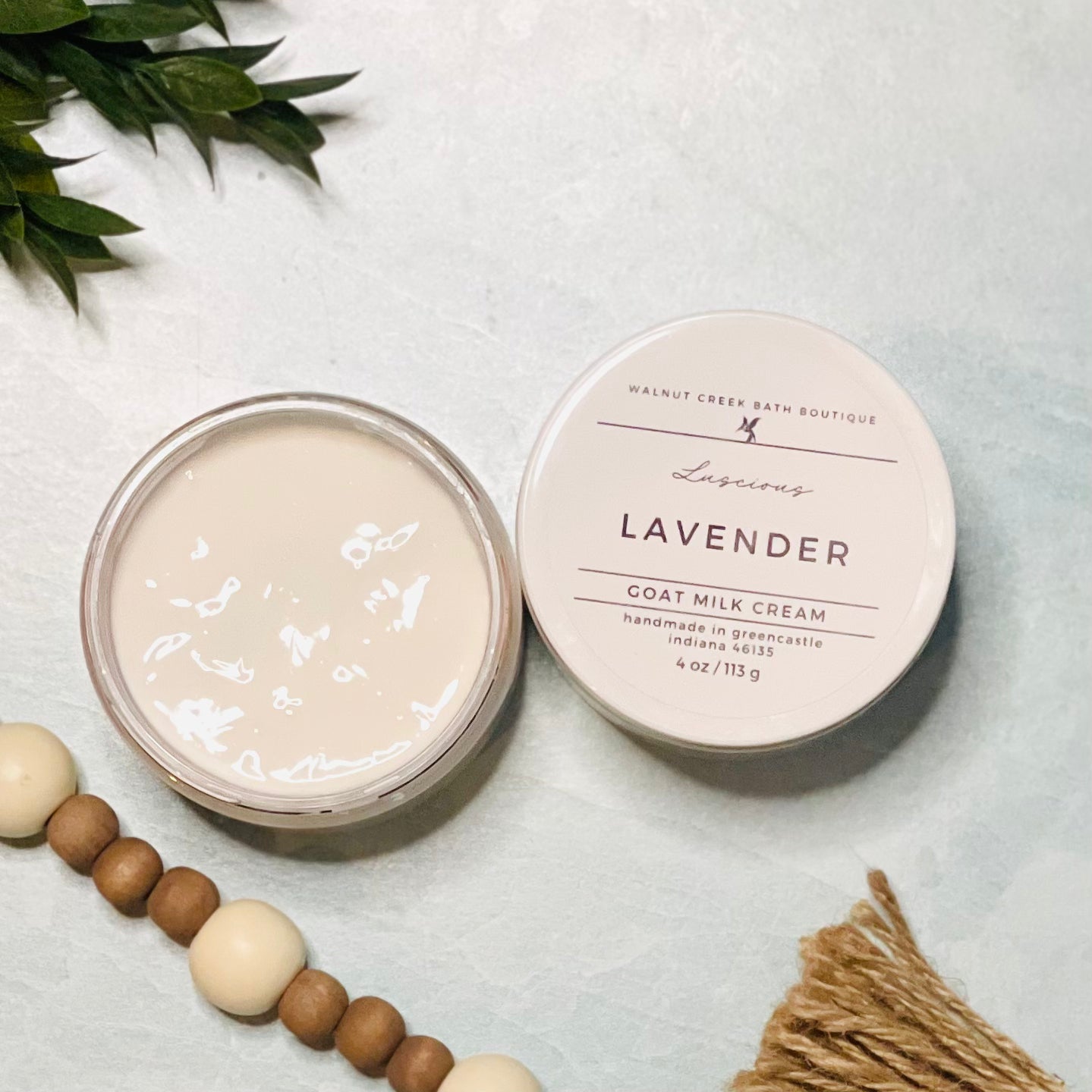 4 oz lavender goat milk cream open with the lid next to it. the texture is so creamy and luscious looking. there is some greenery on the top left and some wooden beads running along the edge with the tassel peaking just in view on the bottom right.