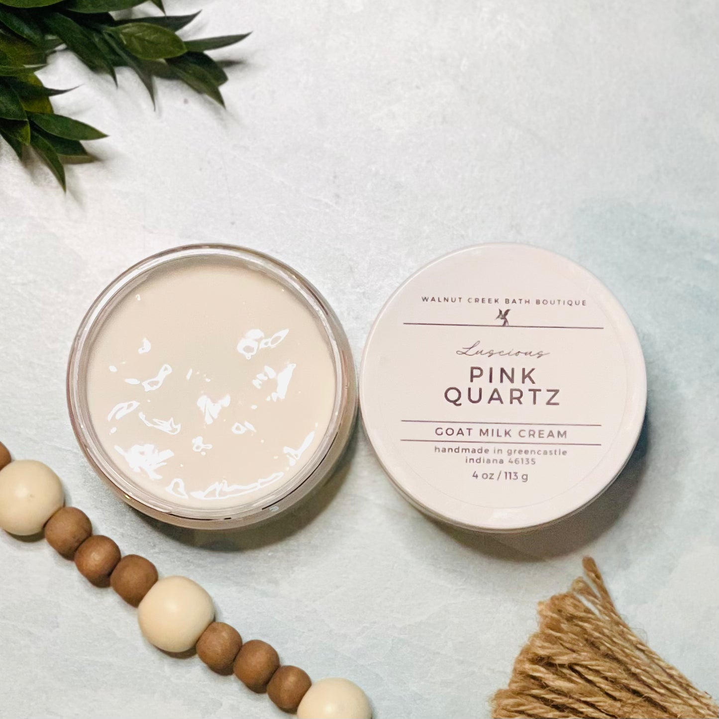 4 oz pink quartz goat milk cream open with the lid next to it. the texture is so creamy and luscious looking. there is some greenery on the top left and some wooden beads running along the edge with the tassel peaking just in view on the bottom right.