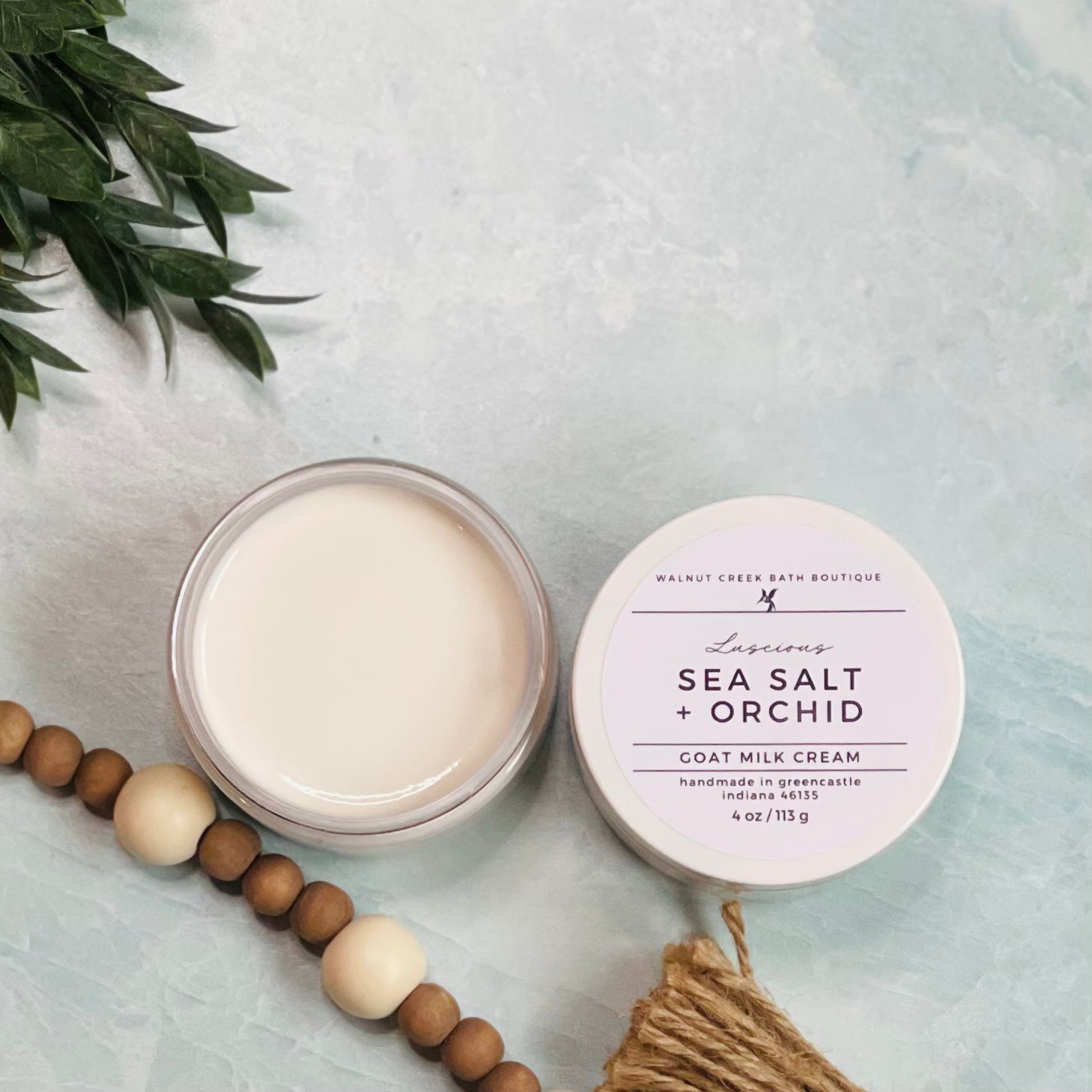 4 oz sea salt and orchid goat milk cream open with the lid next to it. the texture is so creamy and luscious looking. there is some greenery on the top left and some wooden beads running along the edge with the tassel peaking just in view on the bottom right.