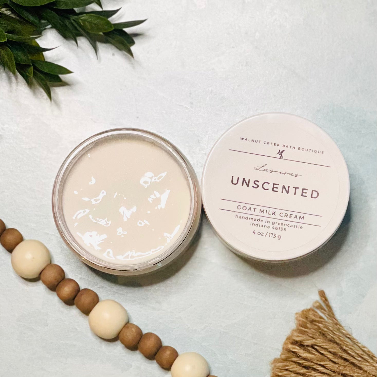 4 oz unscented goat milk cream open with the lid next to it. the texture is so creamy and luscious looking. there is some greenery on the top left and some wooden beads running along the edge with the tassel peaking just in view on the bottom right.