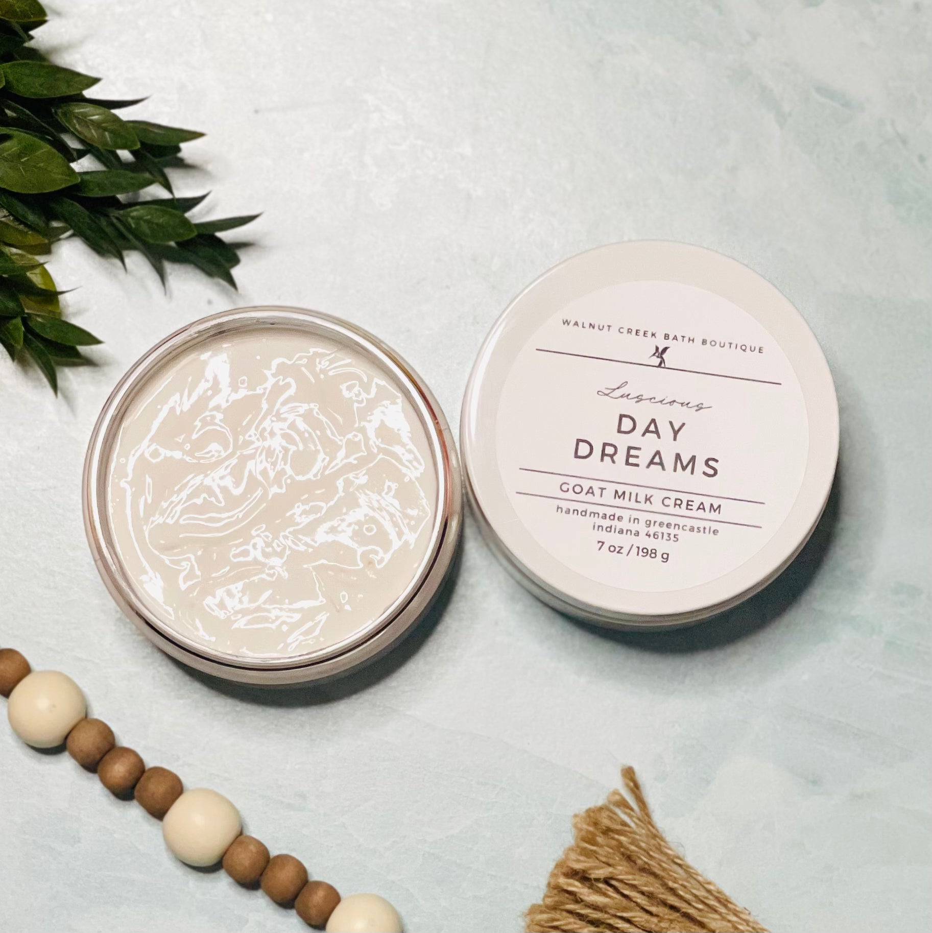7 oz day dreams goat milk cream open with the lid next to it. the texture is so creamy and luscious looking. there is some greenery on the top left and some wooden beads running along the edge with the tassel peaking just in view on the bottom right.