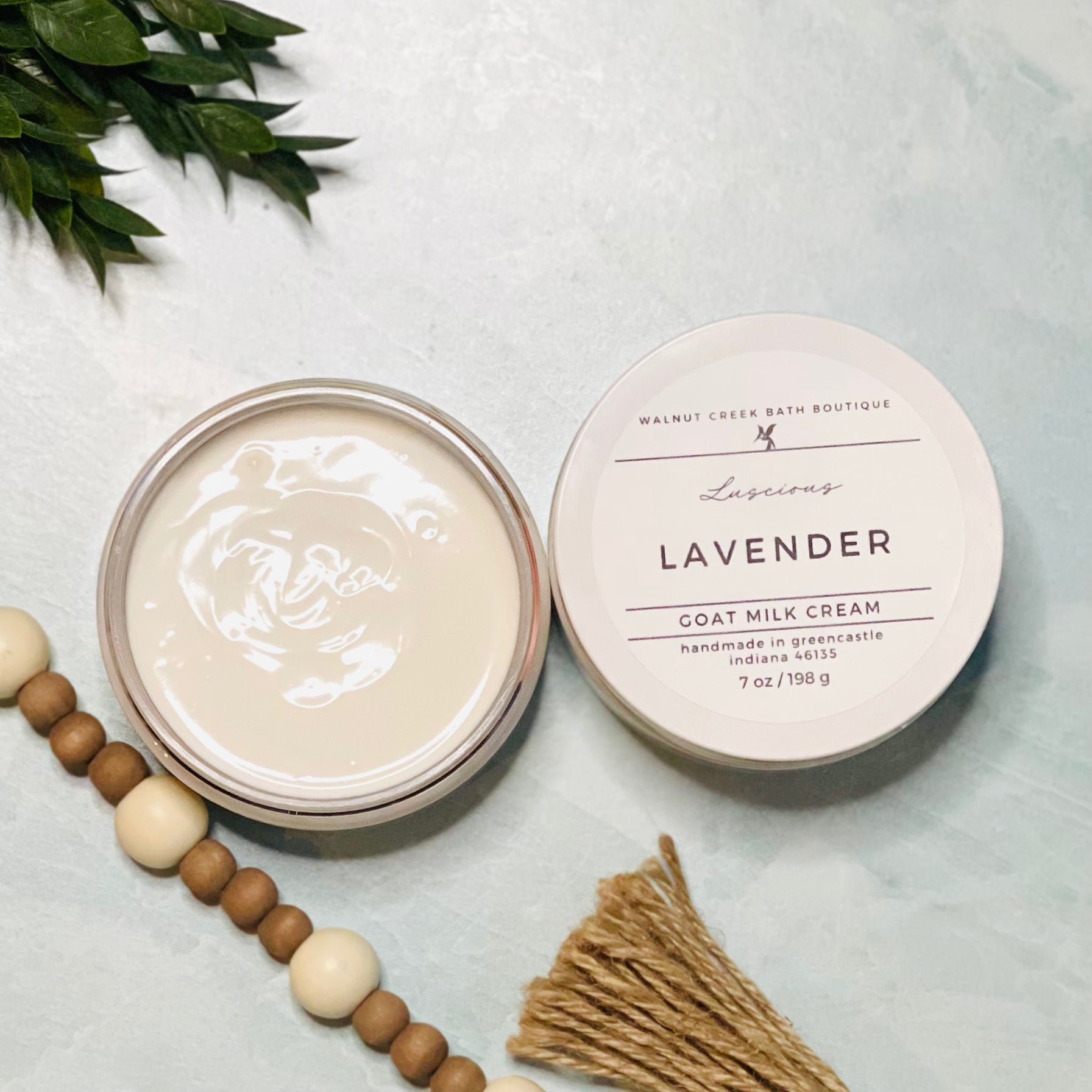 7 oz lavender goat milk cream with the lid next to it. the texture is so creamy and luscious looking. there is some greenery on the top left and some wooden beads running along the edge with the tassel peaking just in view on the bottom right.