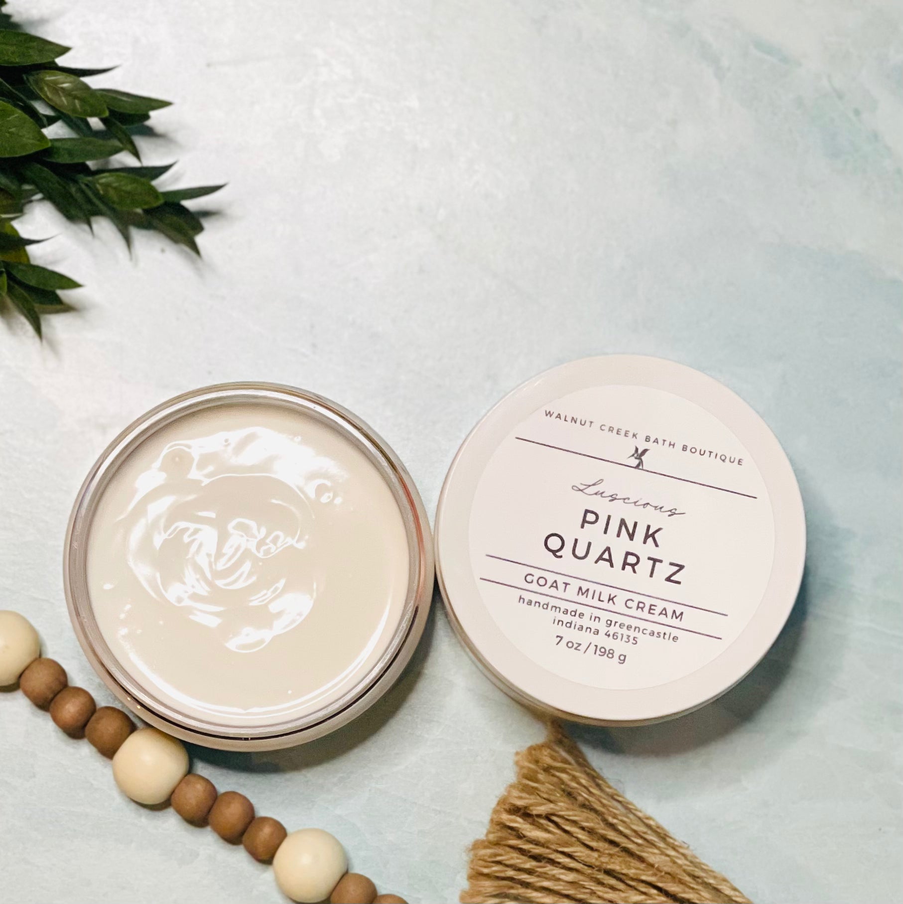7 oz pink quartz goat milk cream open with the lid next to it. the texture is so creamy and luscious looking. there is some greenery on the top left and some wooden beads running along the edge with the tassel peaking just in view on the bottom right.