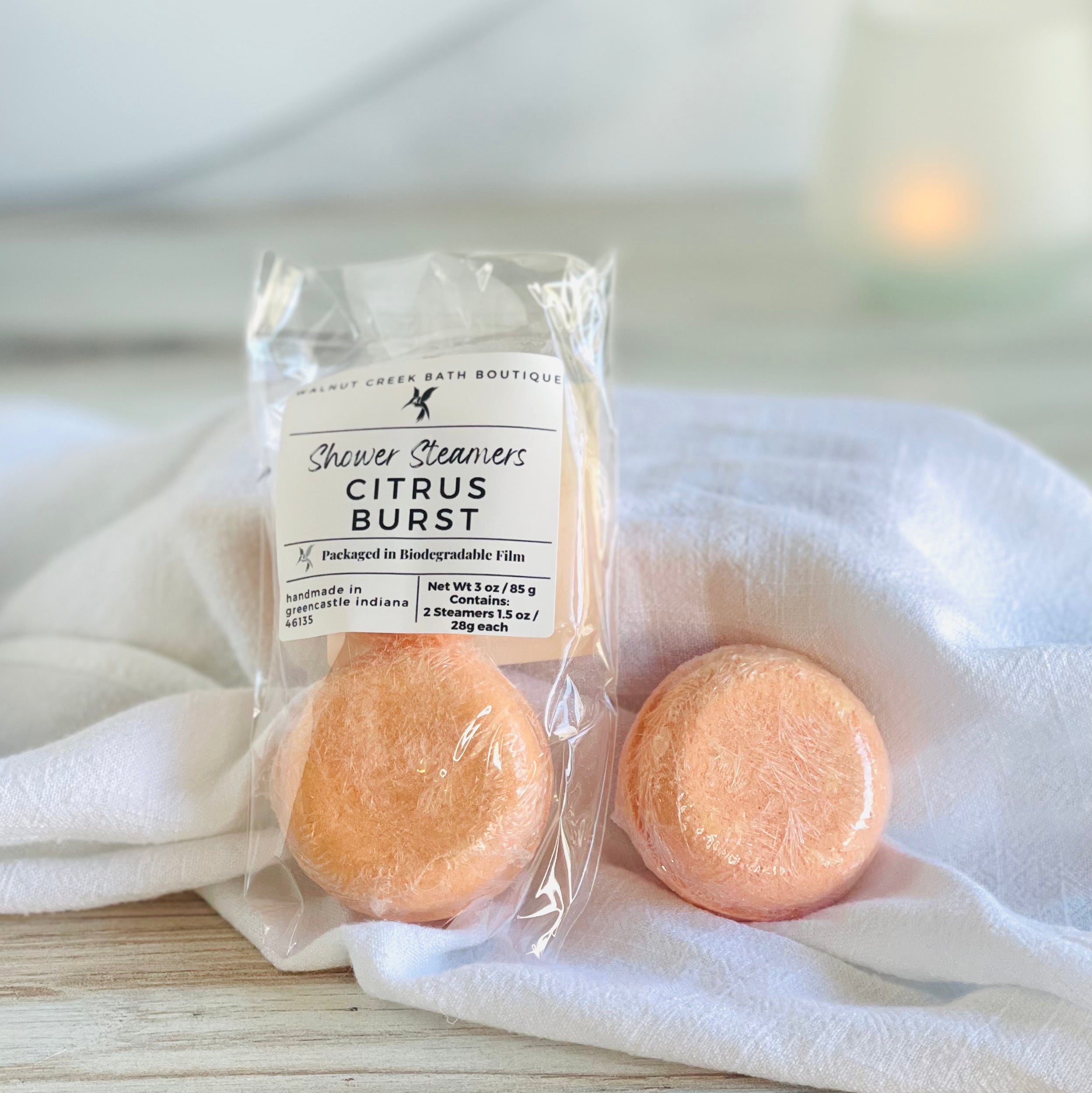 one package of just citrus burst steamers are shown leaning on a white cloth covered box, this shows the pack of 2 and the label. next to this is a single shower steamer still sealed in biodegradable film for freshness.