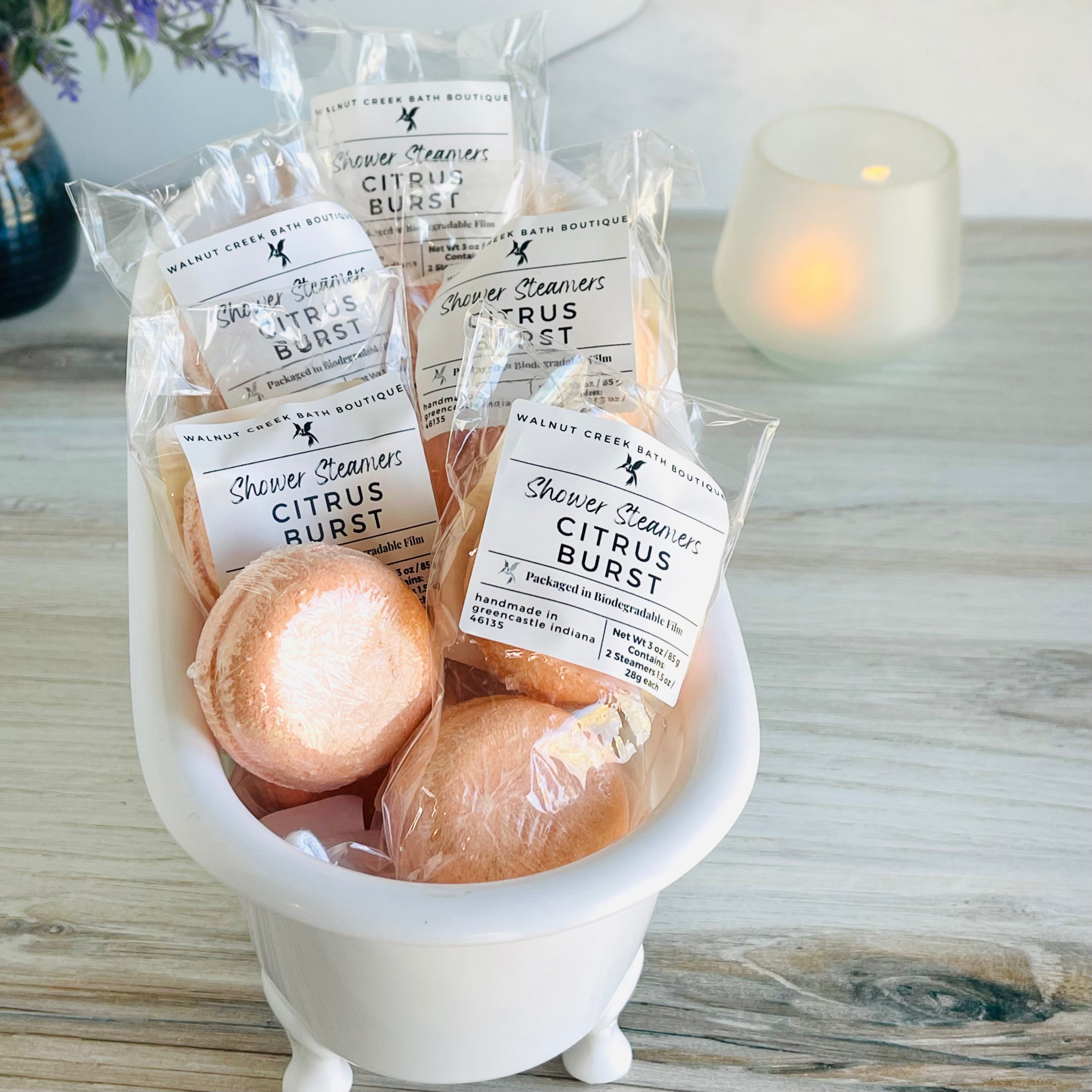 a cute small white tub is brimming with lots of citrus burst shower steamers, with one outside of the package just waiting to be used. in the background is a lit candle to set the mood for a relaxing shower time.