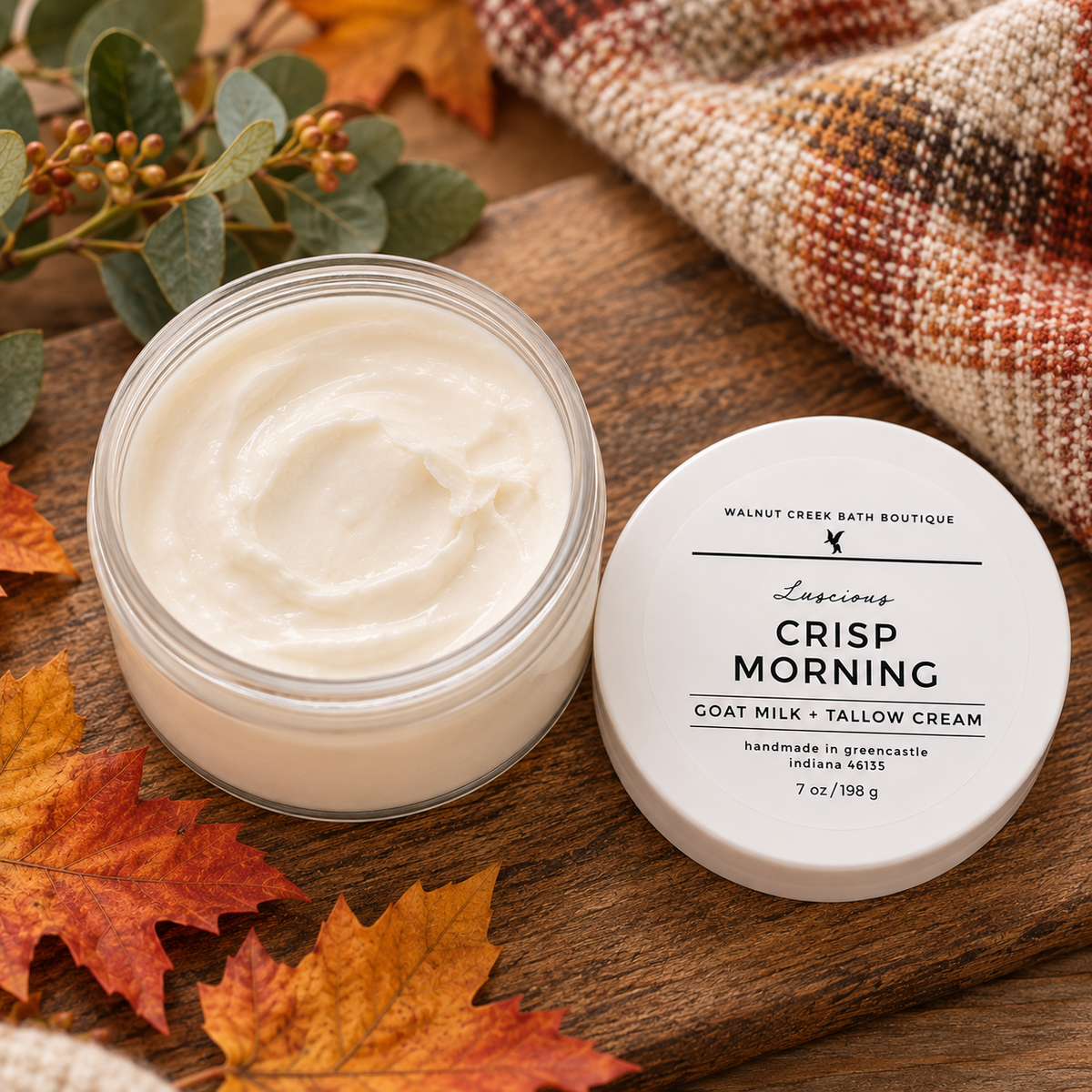 An open jar of Walsh Creek Bath Boutique Lascums Crisp Morning Goat Milk + Tallow Cream sits on a rustic wooden surface next to its white lid. The scene is styled with autumn leaves, greenery, and a plaid blanket.