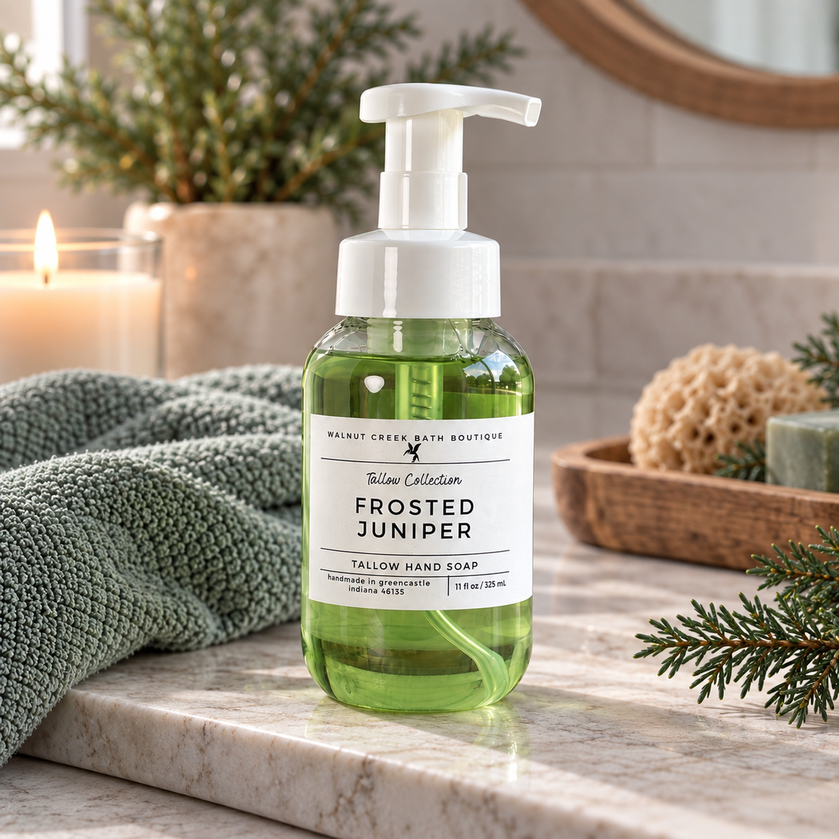 A bottle of Walnut Creek Bath Boutique Frosted Juniper hand soap sits on a marble bathroom counter next to a green towel, with a candle, sponge, and pine branches in the background.