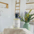 lavender rose micellar water is lifted up on a tan box with some cotton rounds stacked next to it. this is set in front of a spa like bathroom and in the background is a sage green vase holing some greenery
