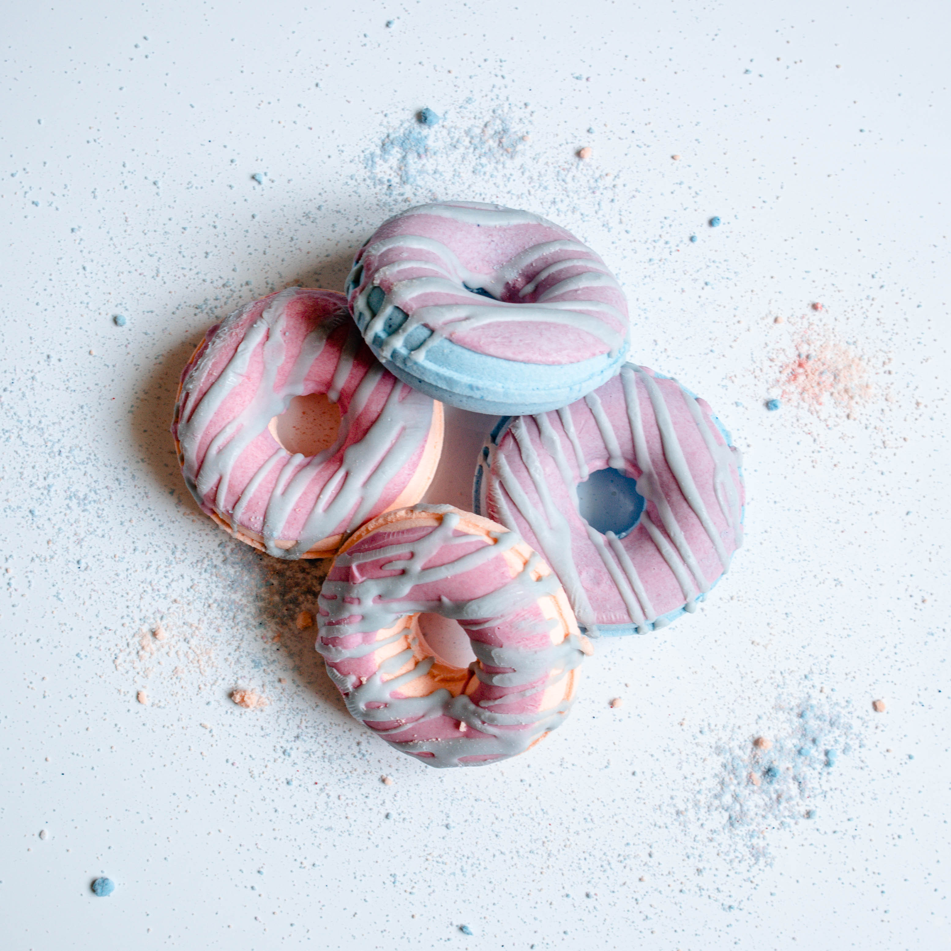 Image shows 4 bath bombs on a white board. The bath bombs are different colors and have icing drizzled on top. there is some bath bomb dust sprinkled around the set for fun interest.