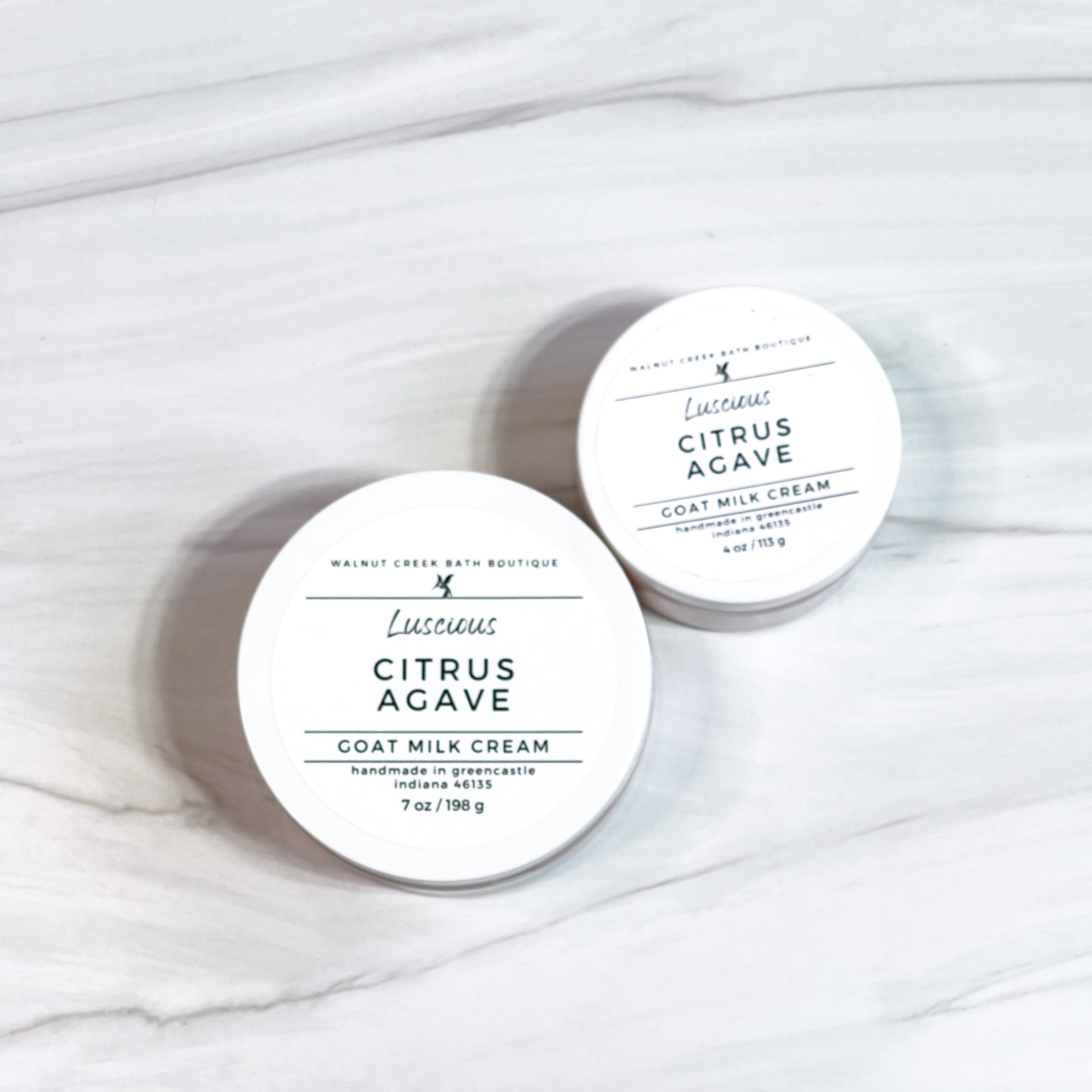 Two round containers of Citrus Agave Goat Milk Cream with white lids, labeled with the product name and branding on a marble surface.