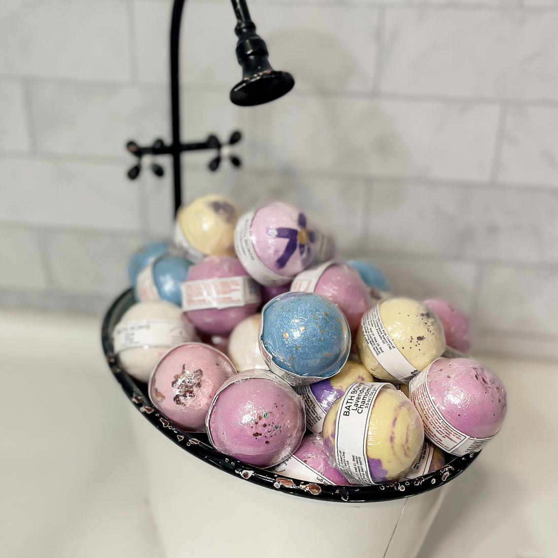 Assorted pastel-colored bath bombs in a black and white metal small tub with a shower in the background.