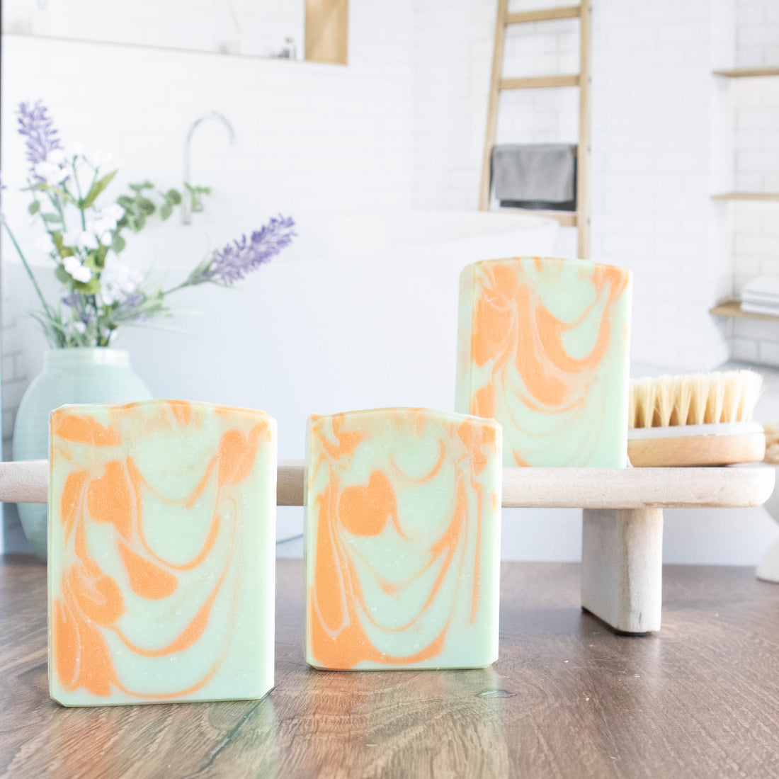 3 agave nectar and lime blossom soaps are shown. they are a light green with an orange swirl. Ons soap is on a wooden stand. there is lavender flowers in a green vase in the background along with a body brush. there is a bathroom scene in the background as well.