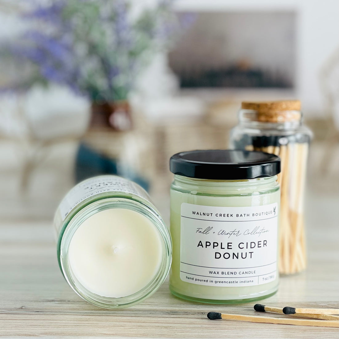 apple cider donut candle with lid to the right of another candle with the top off to show the creamy wax inside. there are a few matches scattered in front and a pretty glass jar of matches in the background