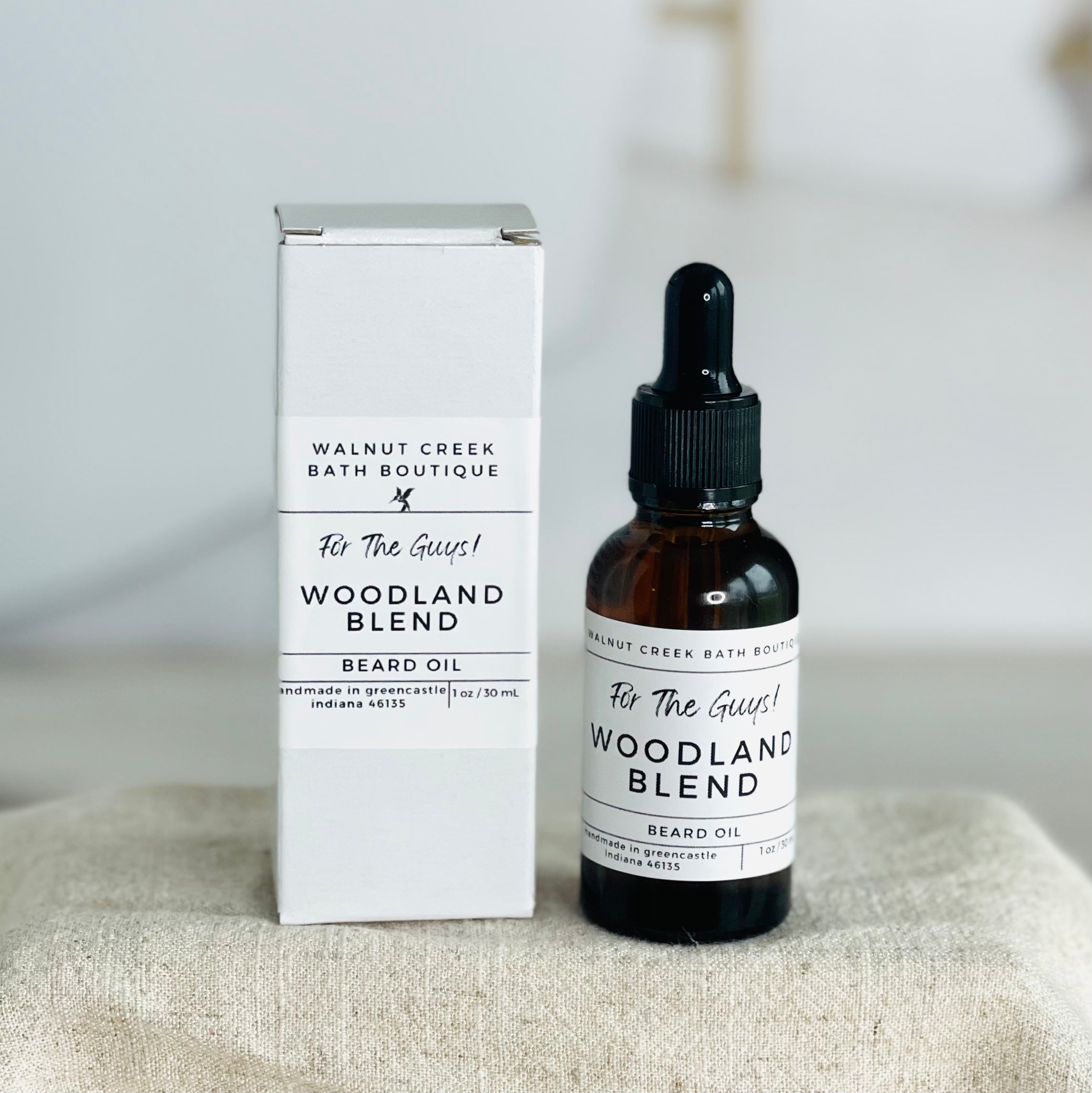 up close look at our beard oil. on the right is the dark amber bottle with the label showing this is a woodland blend scent. on the left is the out box that is white with the same label.