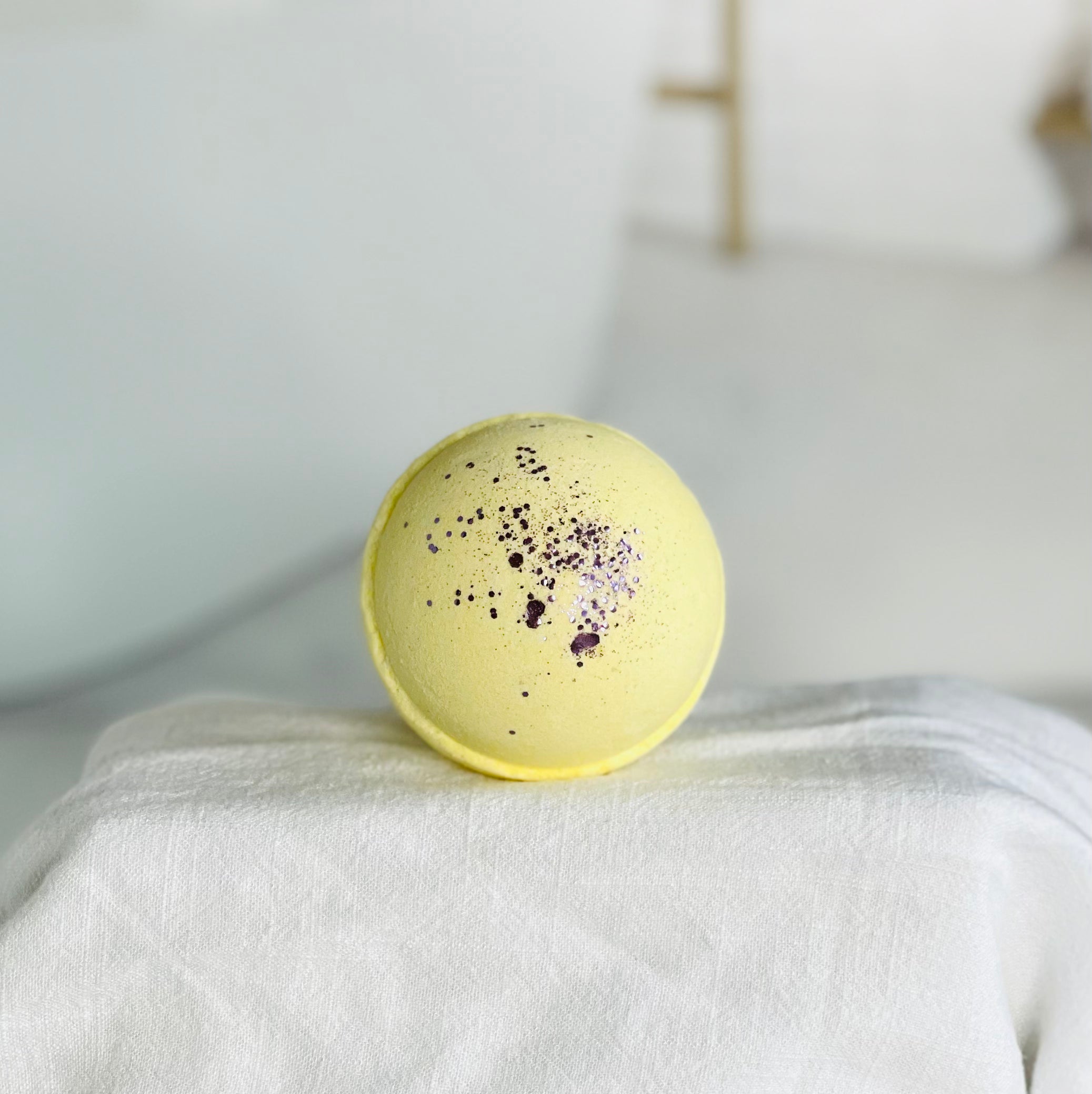 blackberry magnolia bath bomb up close. this is a yellow bath bomb with purple biodegradable glitter. this is sitting center stage on a white cloth over a riser