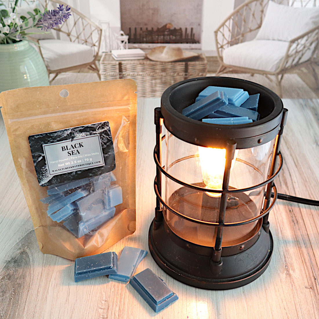 black sea wax melts are light blue and dark blue in color. there is a lit burner filled with these pieces just getting ready to melt. next to this to the left is a bag full of the cute little pieces of waxy goodness!  this is all sitting on a washed out wooden base with a couple of chairs in the background next to a fireplace
