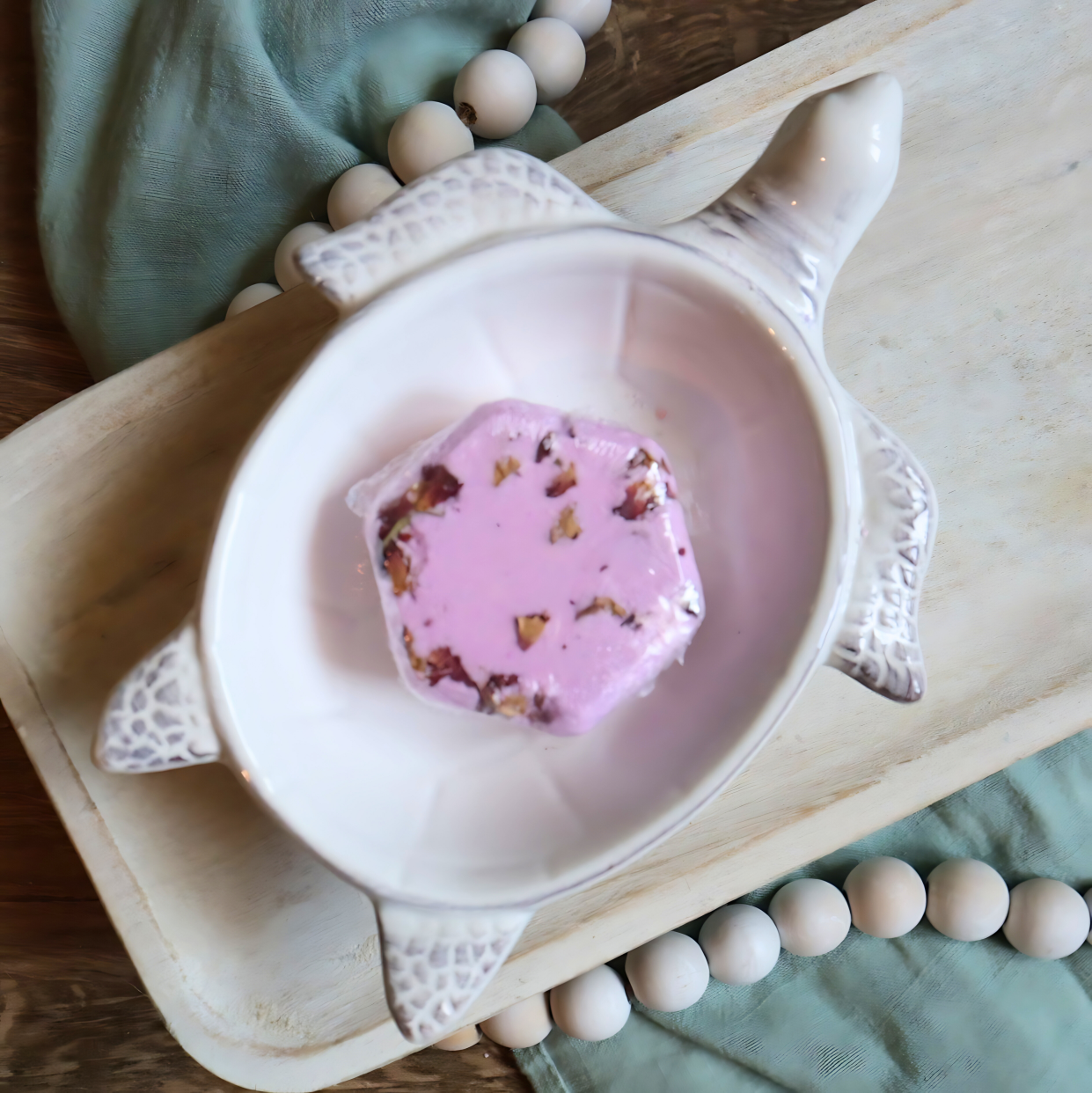 1 bohemian rose bath bomb hexagon is shown in a dish in the shape of a white turtle that is sitting on a tray. the bath bomb is pink and has rose petals over the top. there is a green towel and white beads running beneath the tray