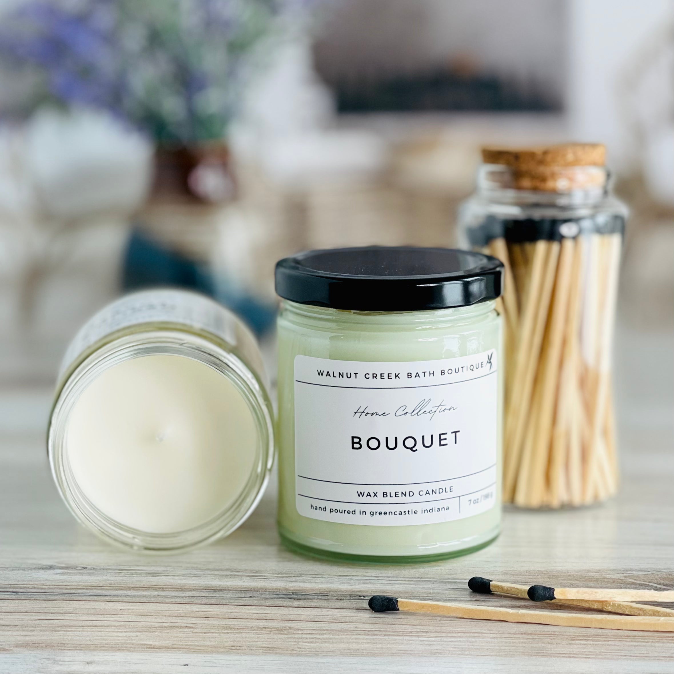 bouquet candle with lid to the right of another candle with the top off to show the creamy wax inside. there are a few matches scattered in front and a pretty glass jar of matches in the background