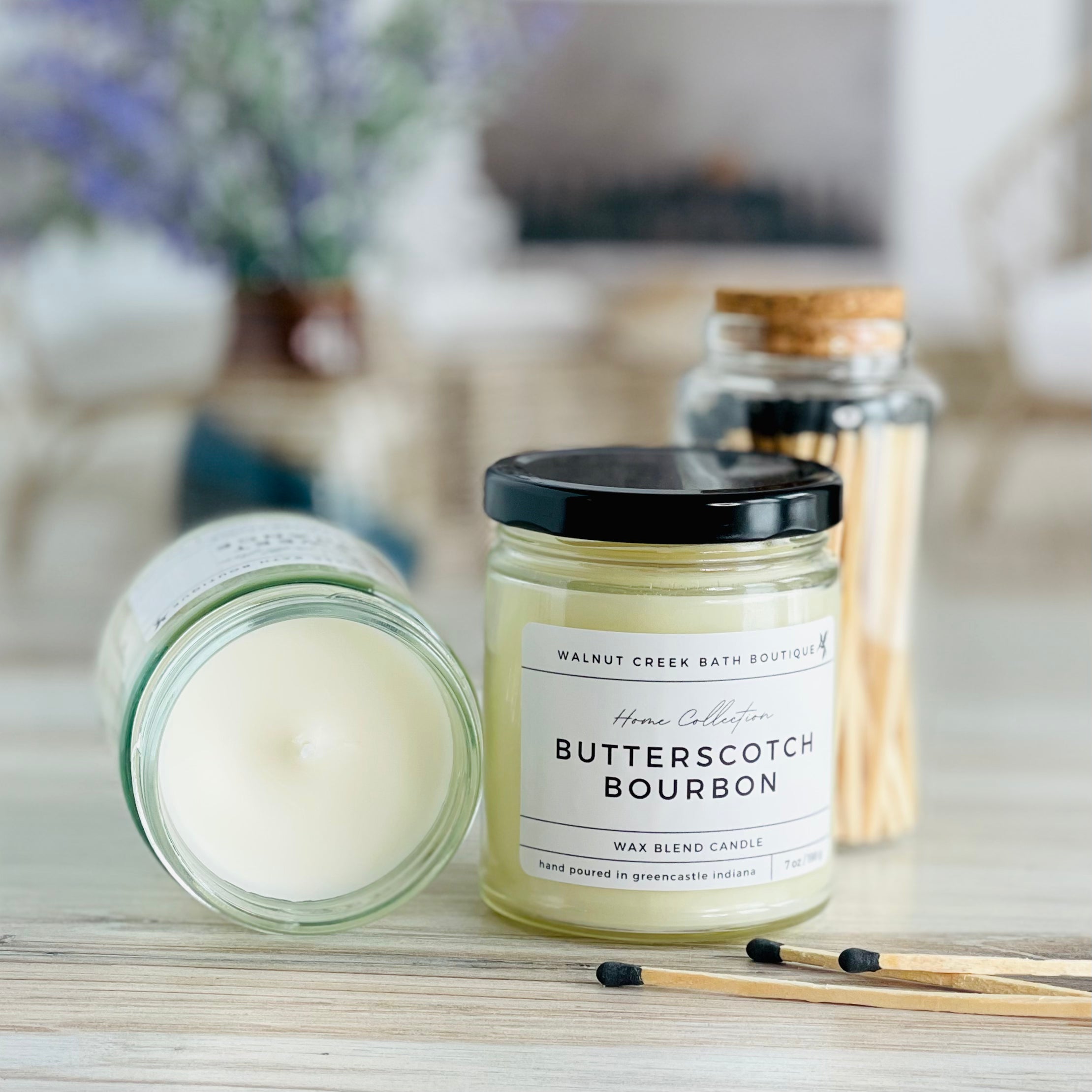 butterscotch bourbon candle with lid to the right of another candle with the top off to show the creamy wax inside. there are a few matches scattered in front and a pretty glass jar of matches in the background