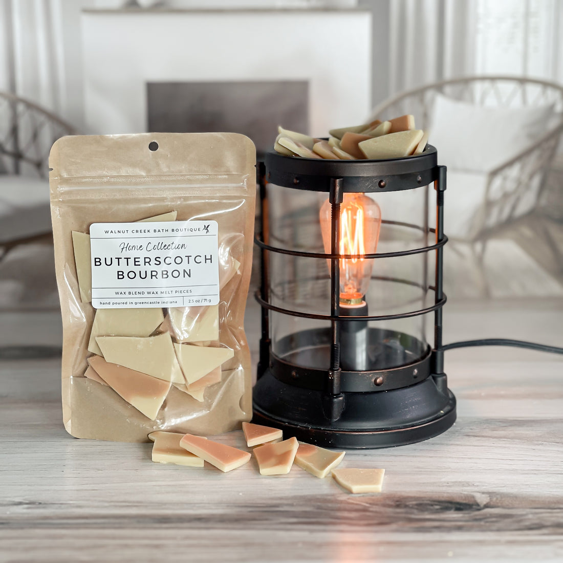 butterscotch bourbon wax melt bag is full and standing next to a rustic farmhouse type burner that is on and filled with the tane brittle pieces of wax. there are also a few shards of the brittle on the table in front of both to show the size and color of the pieces