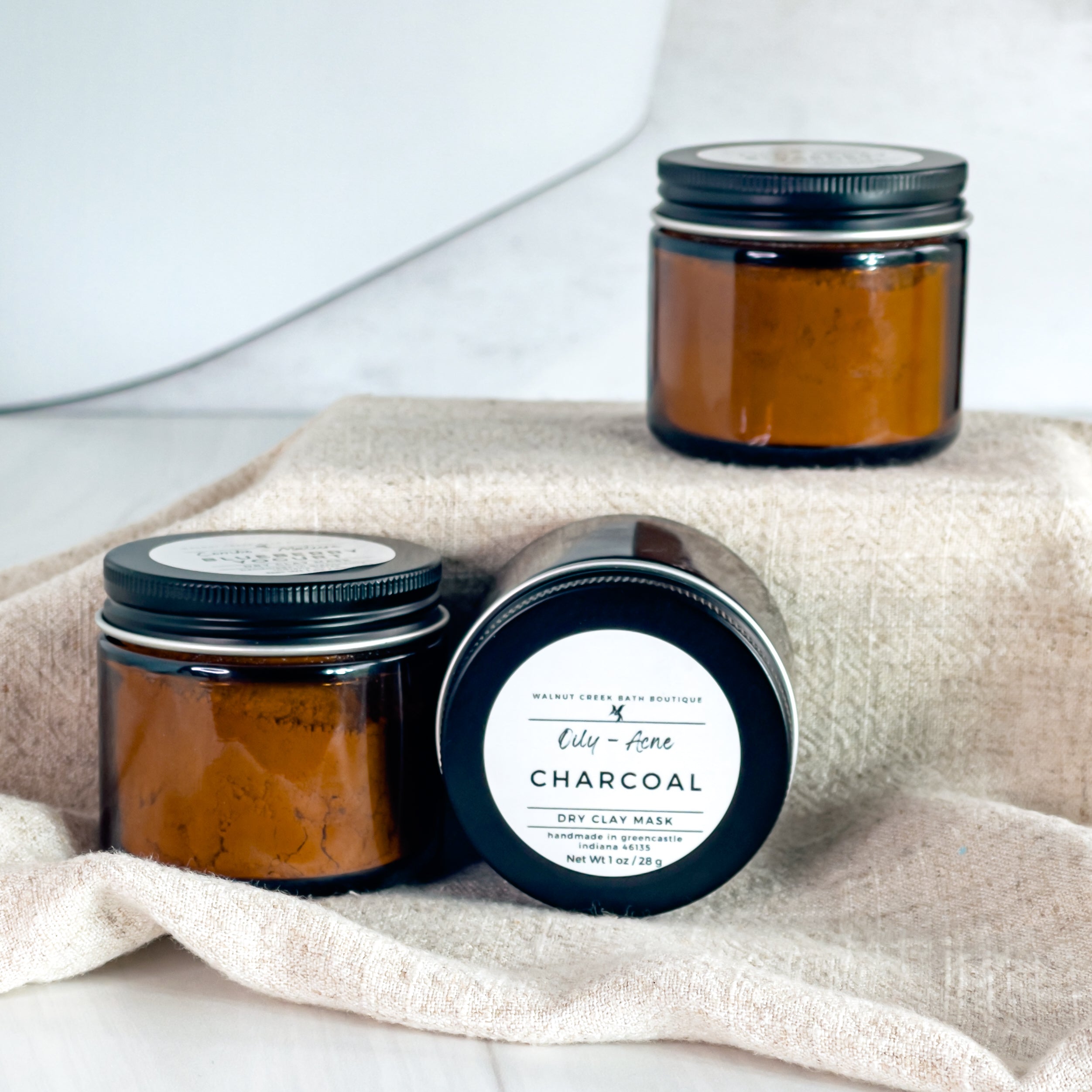 charcoal face masks are shown, one laying on its side to show the simple label while the other two are standing upright. the mask is in amber glass jars with black lids.