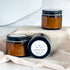 charcoal face masks are shown, one laying on its side to show the simple label while the other two are standing upright. the mask is in amber glass jars with black lids.
