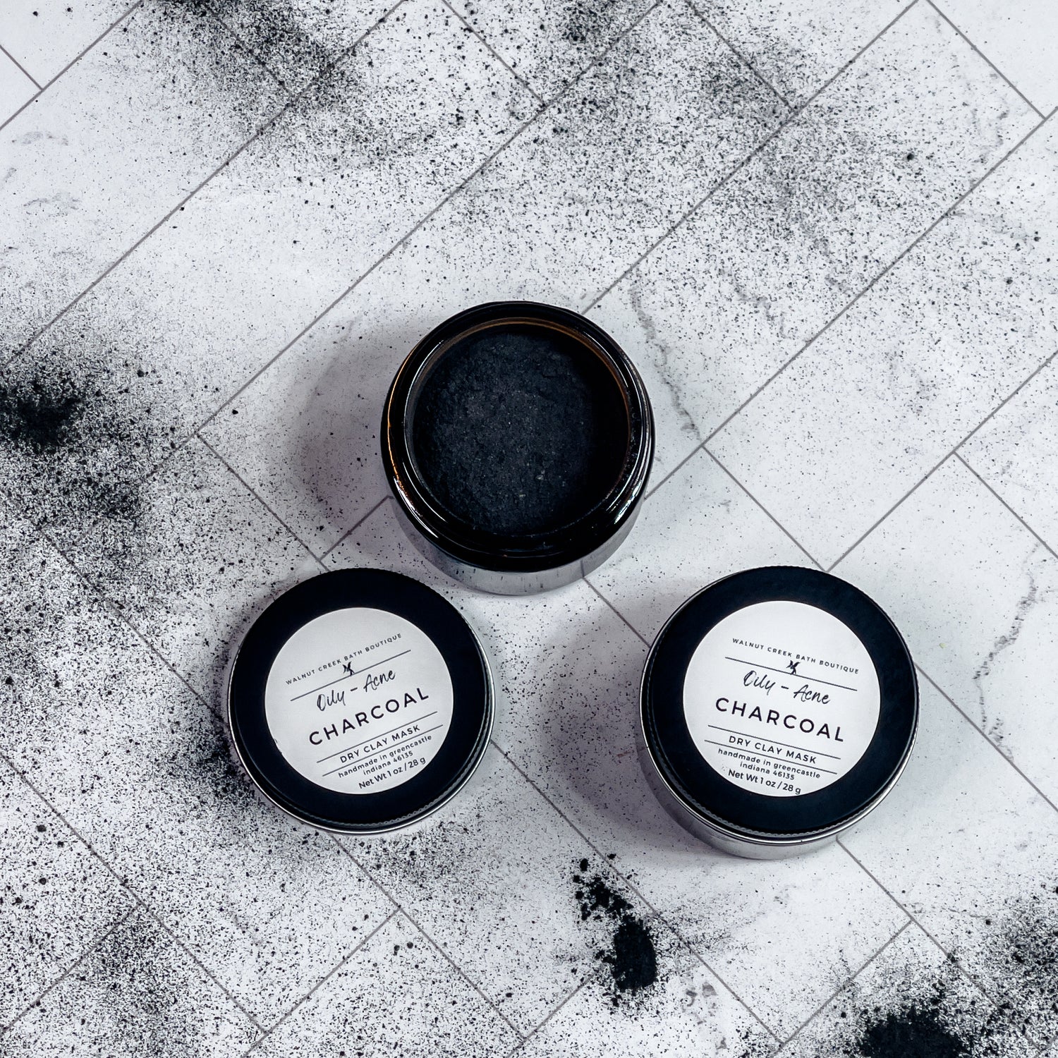 2 charcoal face masks are shown,1 with the top on but one is shown without the top for you to see the black powder inside. there is charcoal powder surrounding these jars , scattered around the surface.