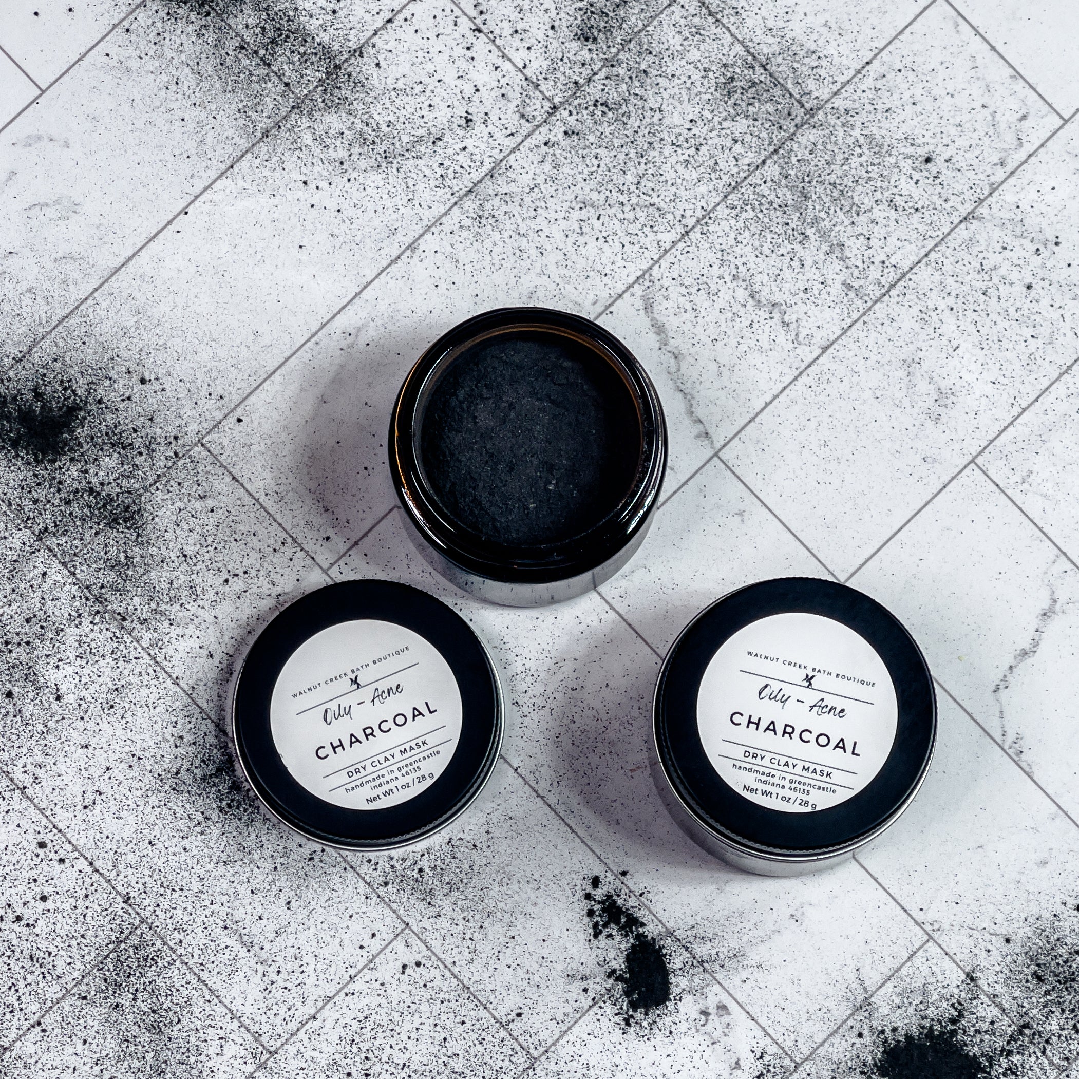 2 charcoal face masks are shown,1 with the top on but one is shown without the top for you to see the black powder inside. there is charcoal powder surrounding these jars , scattered around the surface.