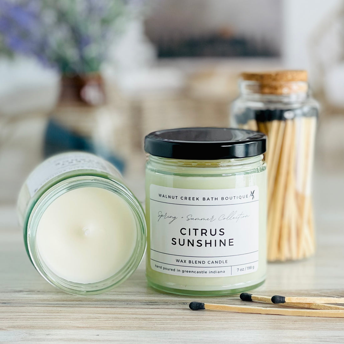 citrus sunshine candle with lid to the right of another candle with the top off to show the creamy wax inside. there are a few matches scattered in front and a pretty glass jar of matches in the background