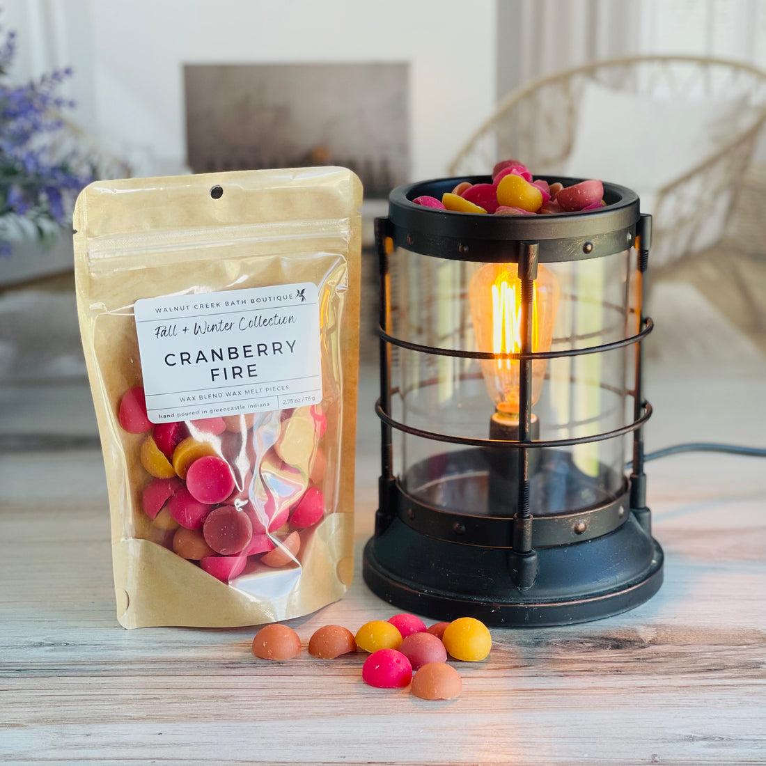 cranberry fire wax melt bag is full and standing next to a rustic farmhouse type burner that is on and filled with the rec, yellow and tan half sphere pieces of wax. there are also a few spheres of the brittle on the table in front of both to show the size and color of the pieces