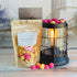 cranberry pumpkin wax melt bag is full and standing next to a rustic farmhouse type burner that is on and filled with the pinks and yellow/orange half sphere pieces of wax. there are also a few spheres of the brittle on the table in front of both to show the size and color of the pieces