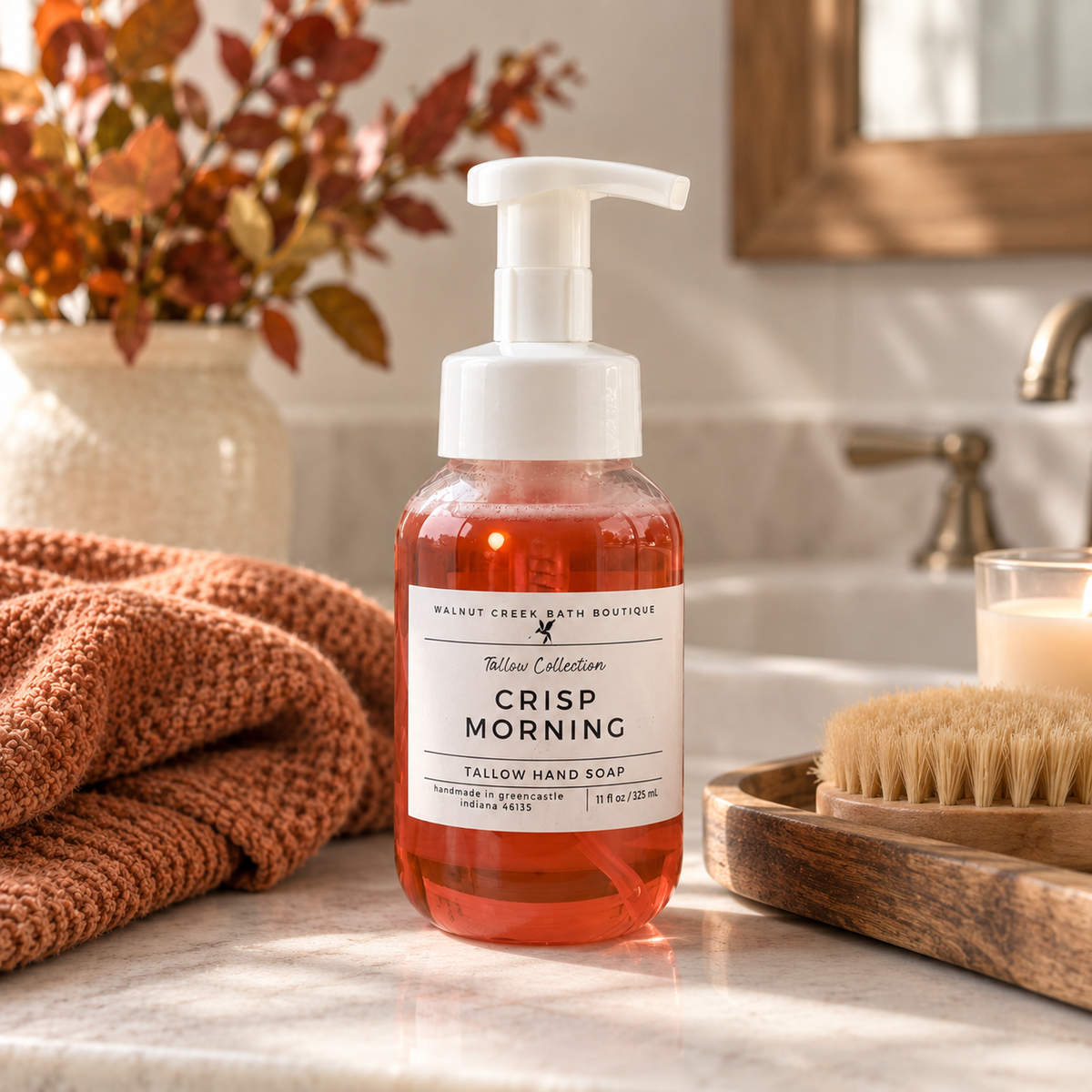 A bottle of Walnut Creek Bath Boutique Thimble Collection Crisp Morning Tallow Hand Soap stands on a marble bathroom counter, surrounded by a rust-colored knit towel, a wooden brush, and a lit candle in a softly blurred bathroom setting.