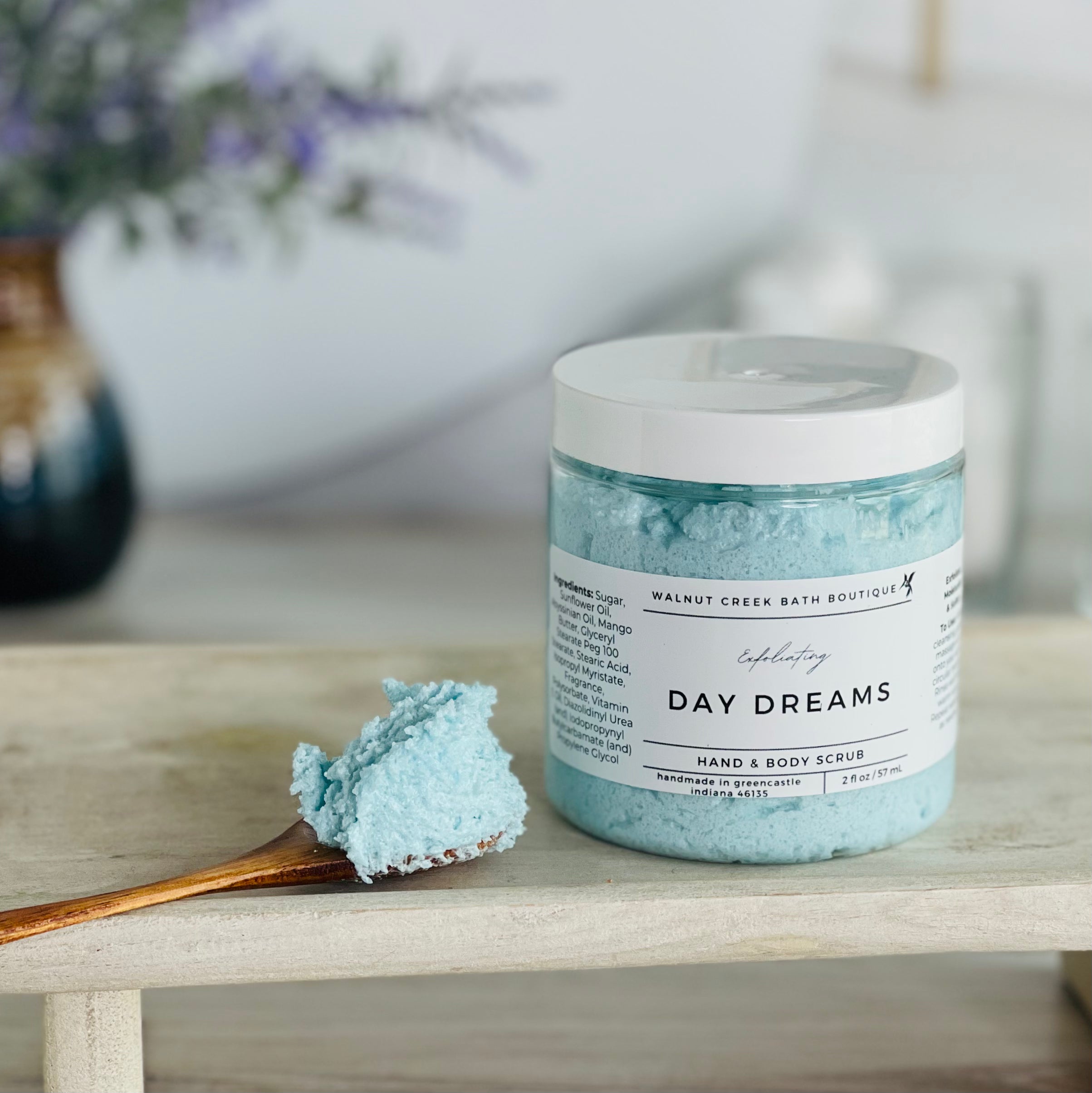 day dreams scrub is shown on a tray with a wooden spoon mounded with the luscious scrub to show this fluffy consistency.. there is some lavender in the background to give interest to this very calm and neutral image to evoke a peaceful experience.