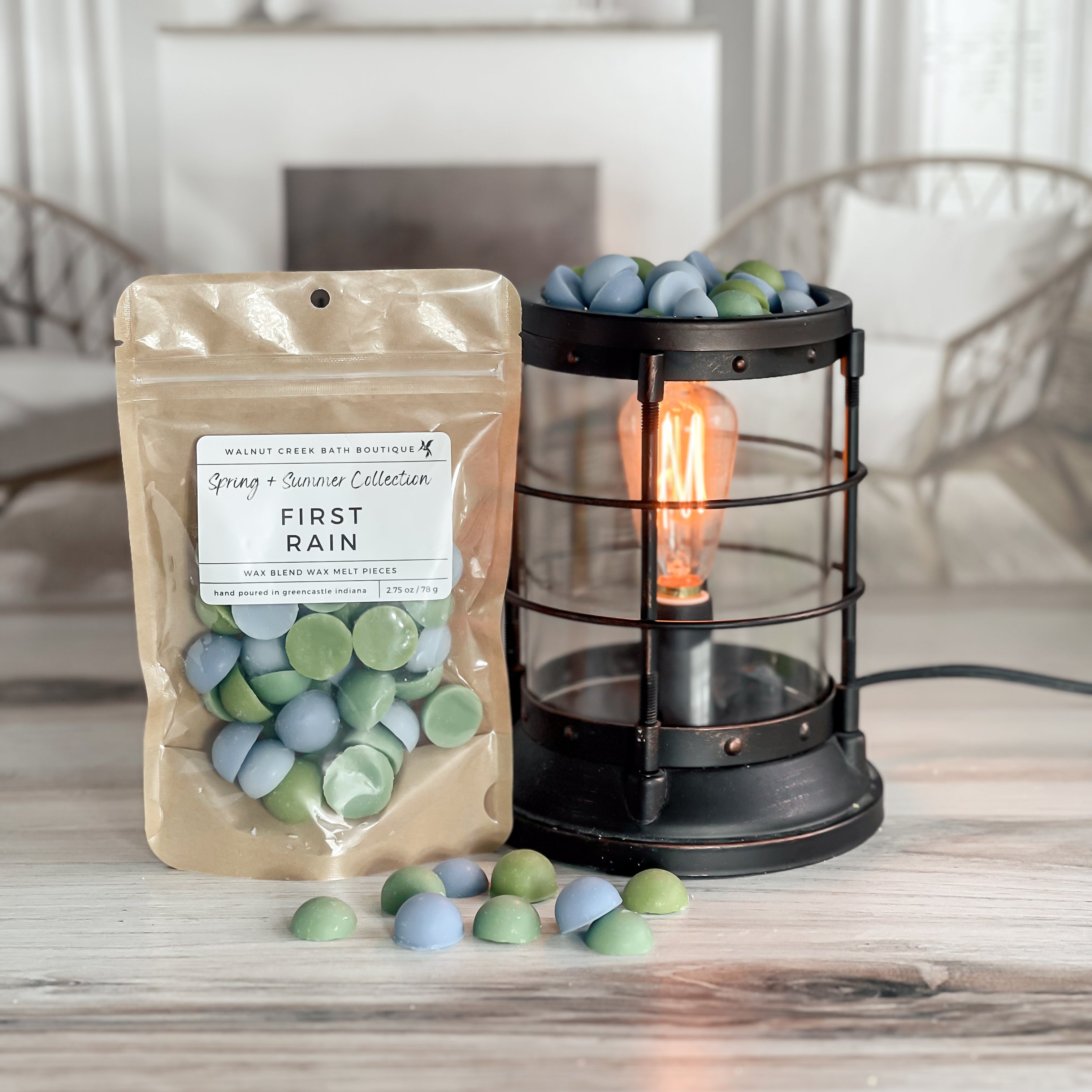 first rain wax melt bag is full and standing next to a rustic farmhouse type burner that is on and filled with the green and blue half sphere pieces of wax. there are also a few rounds of the wax on the table in front of both to show the size and color of the pieces