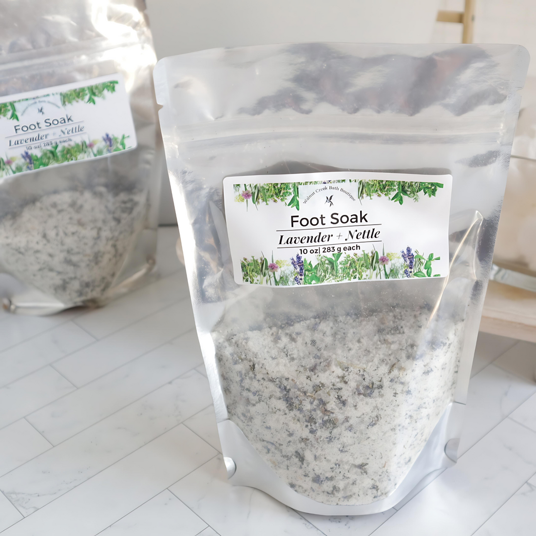 a large bag of foot soak is shown with botanicals on the label and a window to show all the salts and botanicals in the bag. there is another bag sort of in the background to the left.