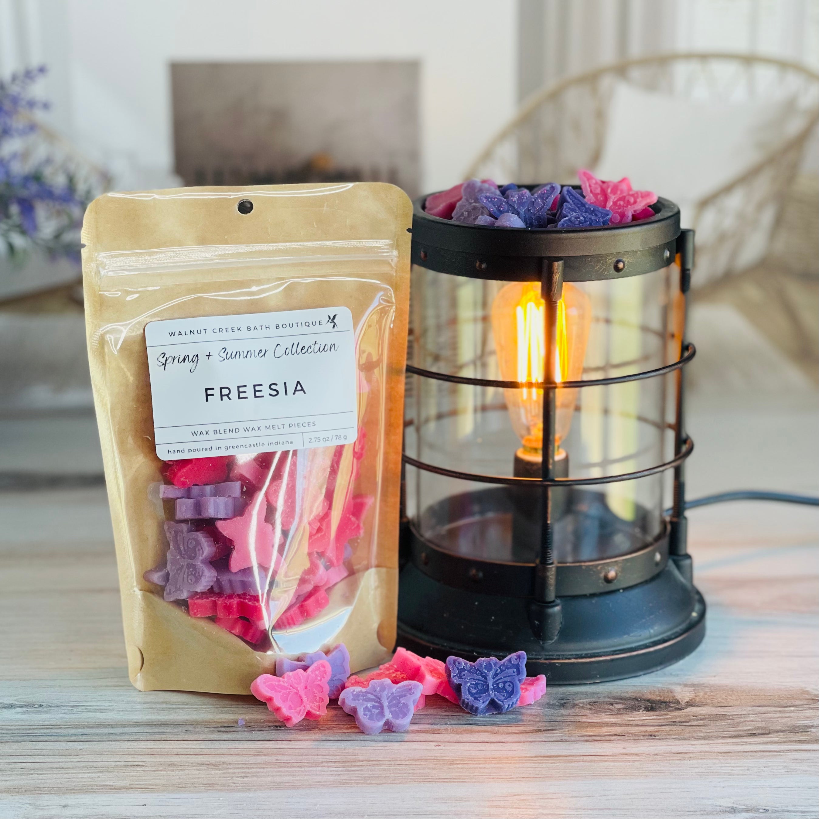 freesia wax melt bag is full and standing next to a rustic farmhouse type burner that is on and filled with the pinks and purples butterfly shapes of wax. there are also a few butterflies on the table in front of both to show the size and color of the pieces
