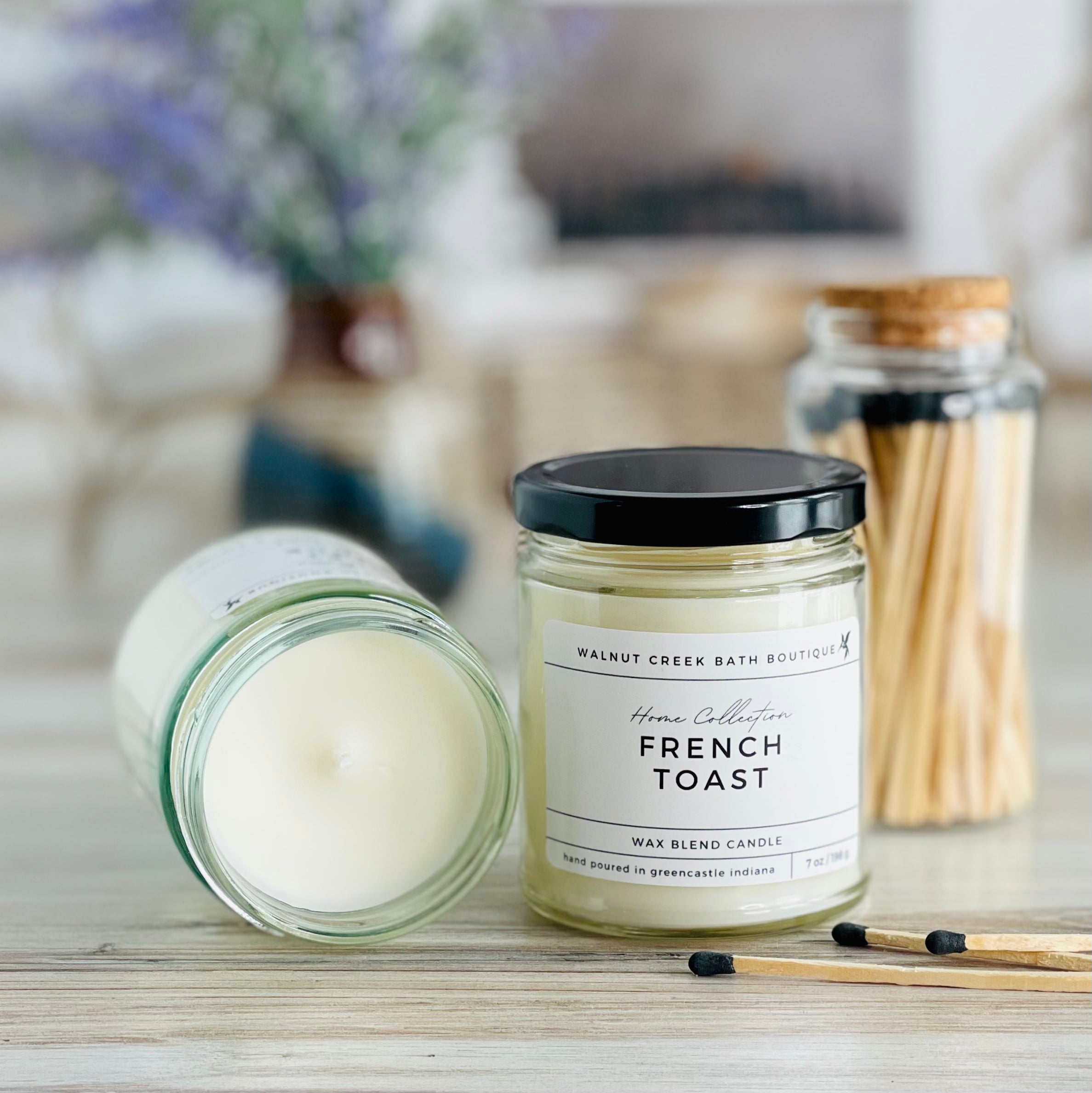french toast candle with lid to the right of another candle with the top off to show the creamy wax inside. there are a few matches scattered in front and a pretty glass jar of matches in the background