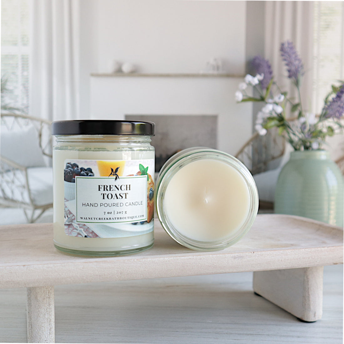 on french toast candle is sitting on a riser with the label forward and the black top on. next to this is an open candle laying flat to show the smooth top of the candle. in the background is a green vase of lavender buds and a fireplace flanked by a couple of wicker chairs.