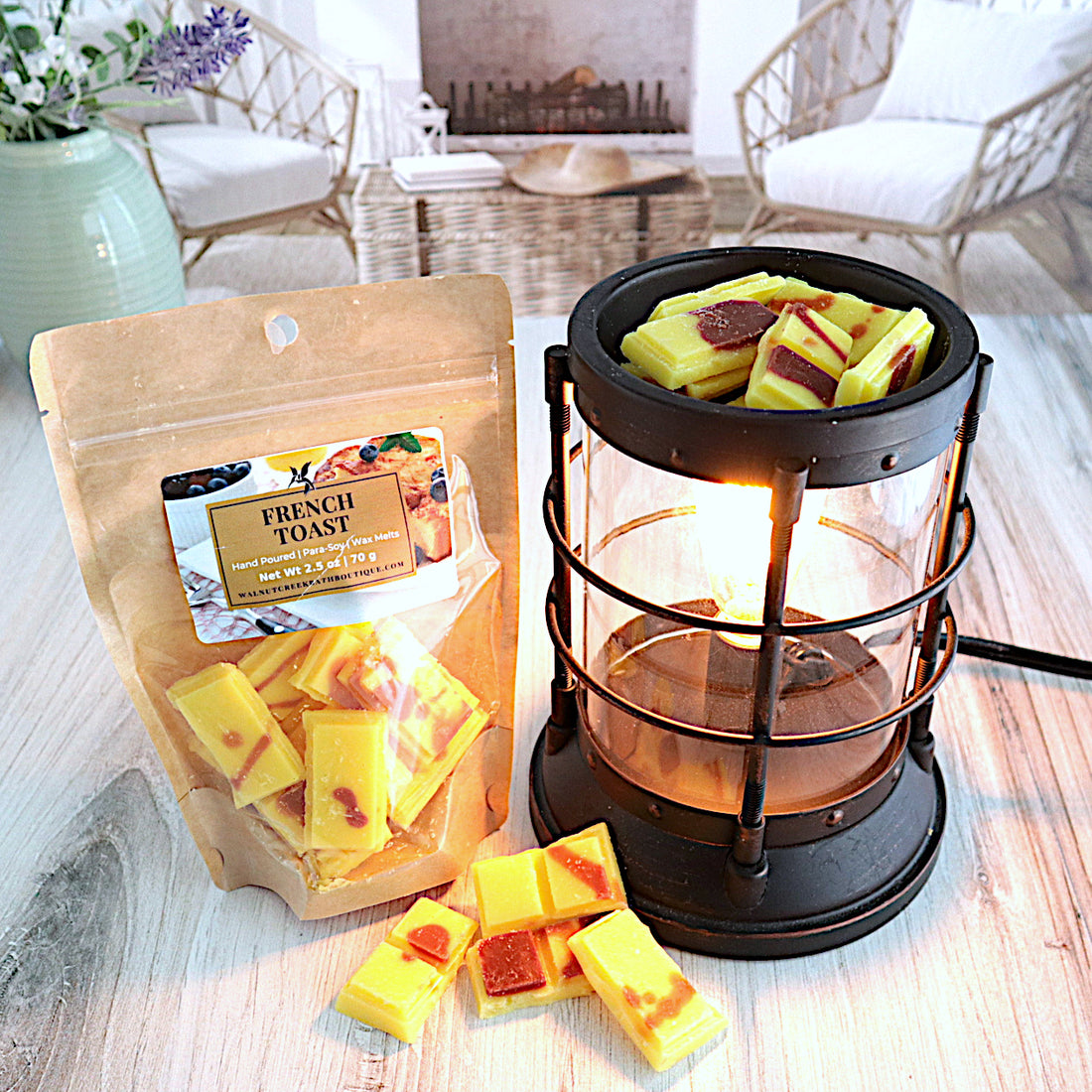 a bag of french toast wax melts are standing next to a lit burner with some of the wax pieces in the cup. there are more wax melt pieces on the table between the bag and the burner. the pieces are yellow in color with some caramel colored drizzles.  this is all sitting on a washed out wooden base with a couple of chairs in the background next to a fireplace 