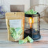 frosted juniper wax melt bag is full and standing next to a rustic farmhouse type burner that is on and filled with the green brittle pieces of wax. there are also a few shards of the brittle on the table in front of both to show the size and color of the pieces
