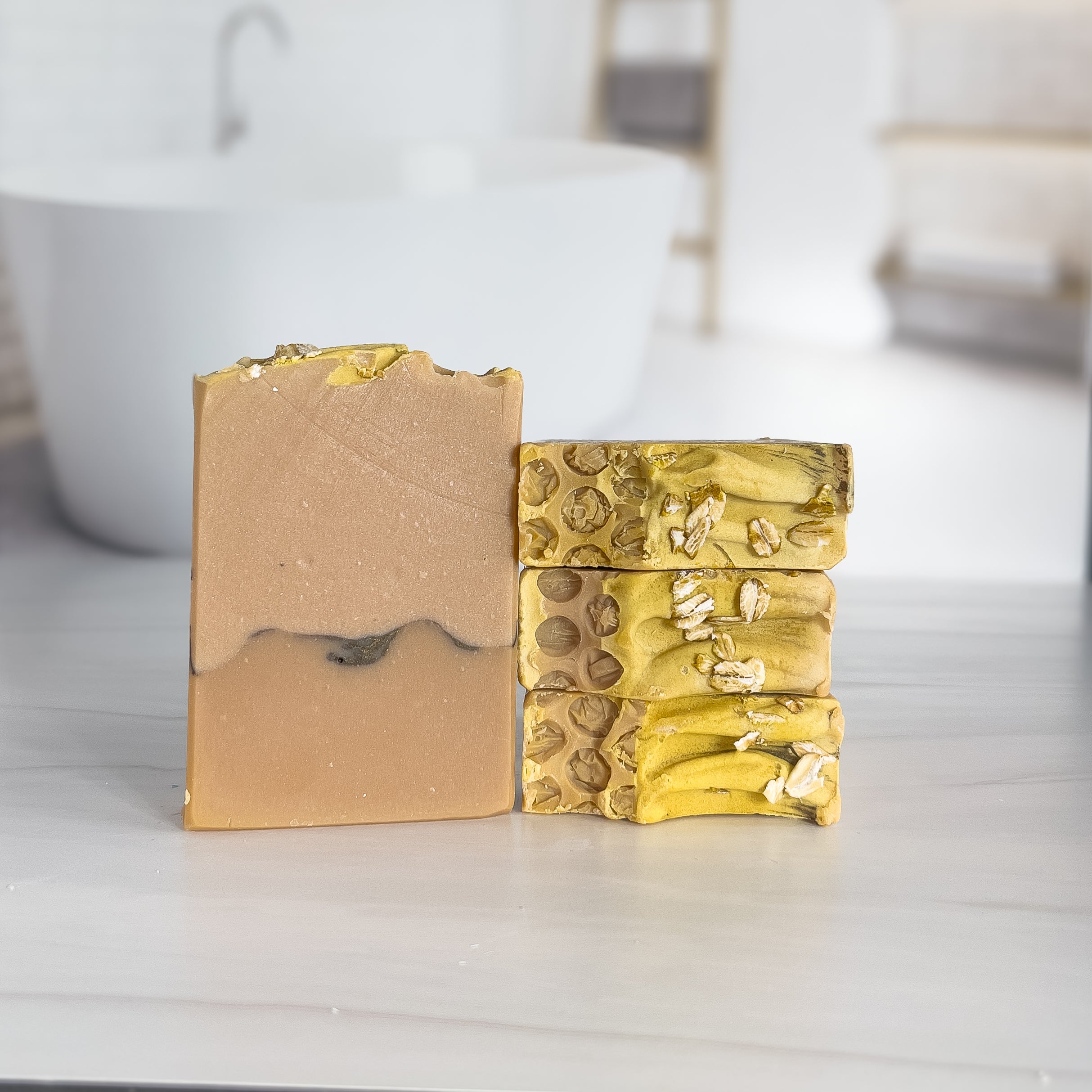 there are golden oat soaps stacked to show the cute honeycomb texture on the tops, along with a sprinkling of oats. next to these is another bar to show the darker bottom and the dark line and then on top of that a lighter honey color.
