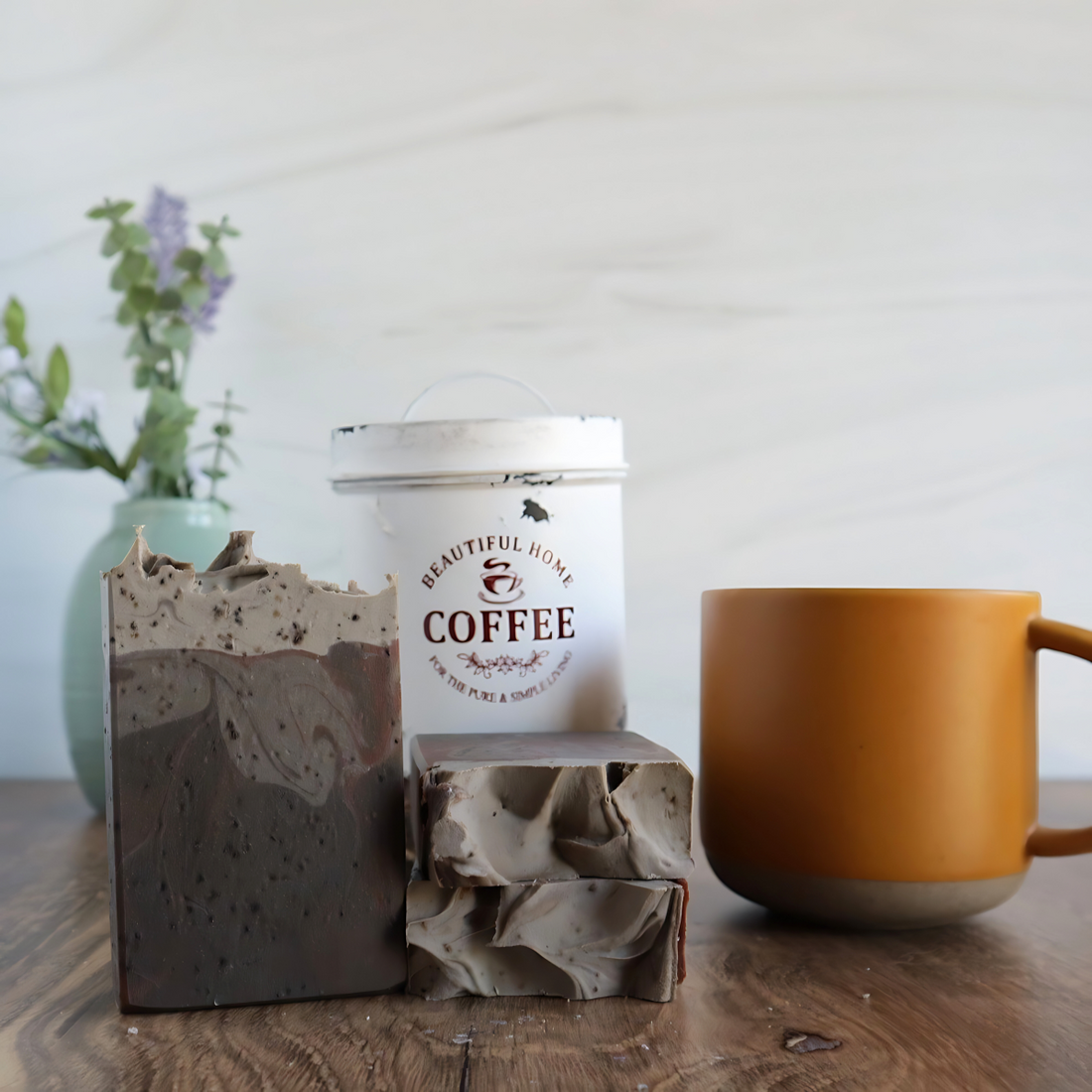 3 hazelnut coffee soaps are shown, one standing facing you to show the dark to light swirls and specs of real coffee grounds throughout. 2 more are laying flat to show a sculpted top. to the right of these is a coral colored coffee cup and in the background there is an antique white tin with the words coffee on it.