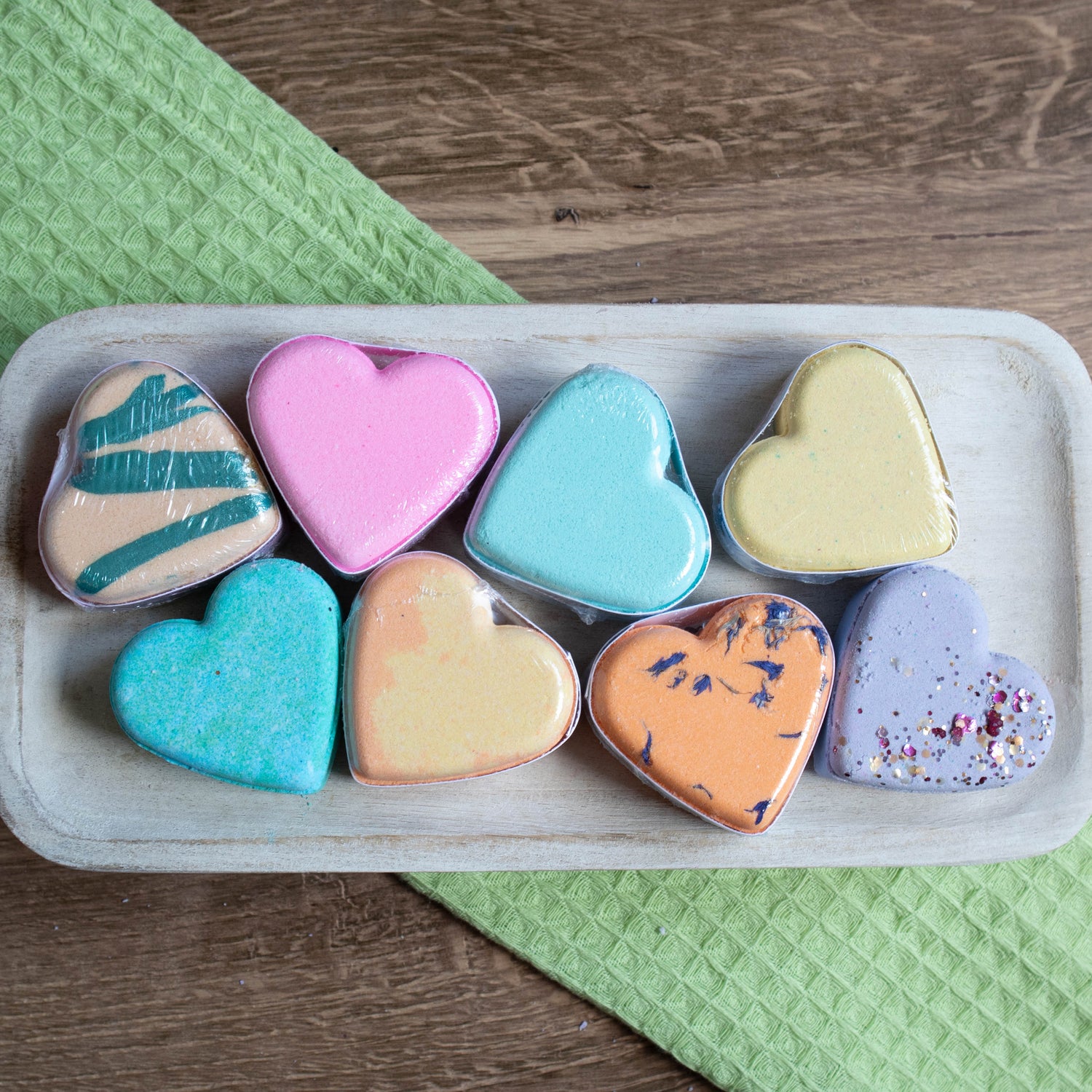 a bunch of different colored heart bath bombs are scattered on a wooden tray. some have stripes of colors, one is orange with blue flowers, one is a lavender color with pink and gold glitter.