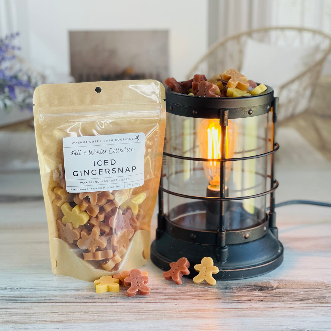 iced gingersnap wax melt bag is full and standing next to a rustic farmhouse type burner that is on and filled with the tan and brown gingerbread men pieces of wax. there are also a few gingerbread men on the table in front of both to show the size and color of the pieces