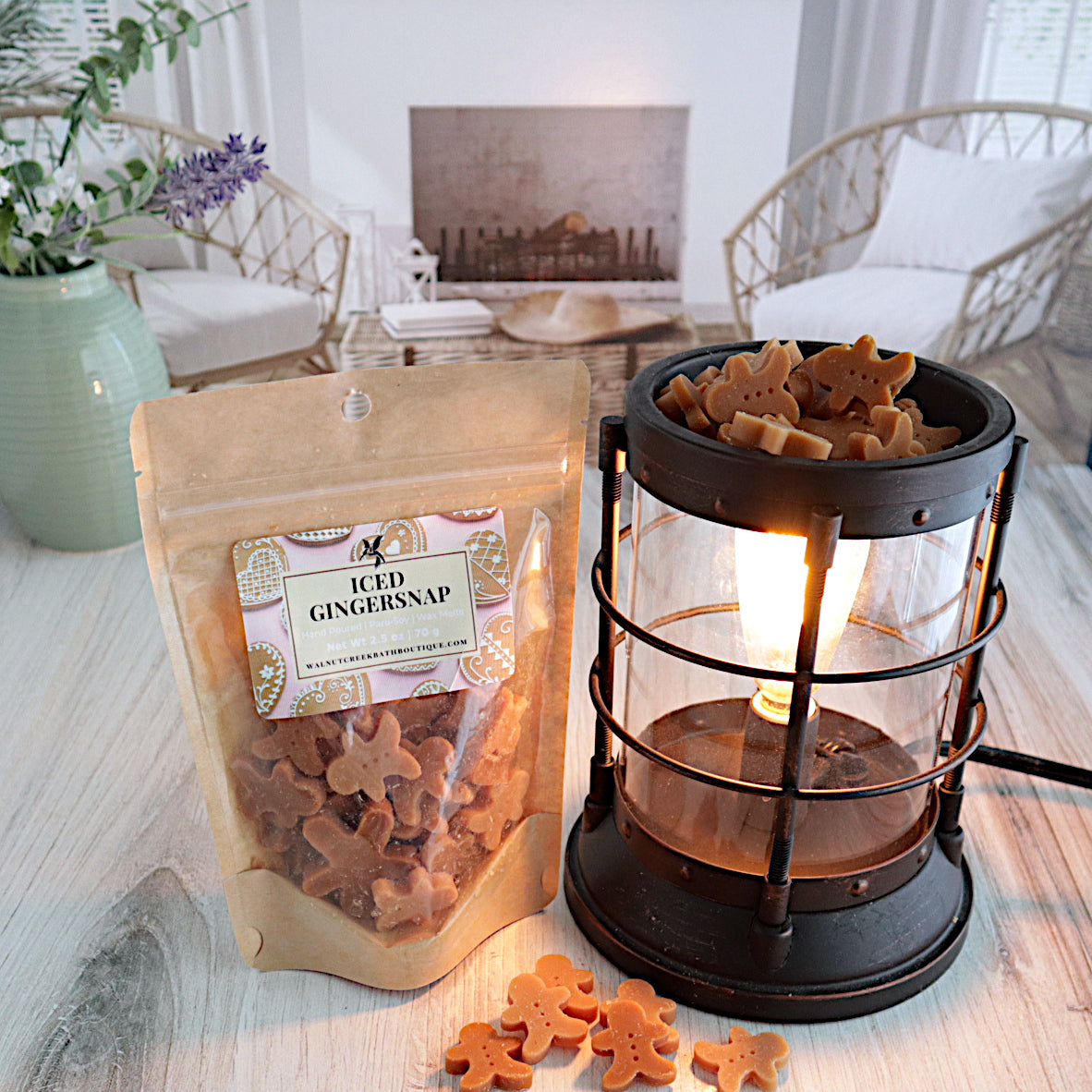 a bag of iced gingersnap wax melts is standing next to a lit burner that is full of the wax pieces. there is a scattering of the wax pieces on the table in front of the burner. the wax is in the shape of light and dark brown gingerbread men. this is all sitting on a washed out wooden base with a couple of chairs in the background next to a fireplace
