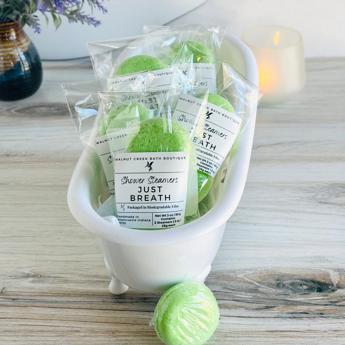 a cute small white tub is brimming with lots of just breath shower steamers, with one outside of the package just waiting to be used. in the background is a lit candle to set the mood for a relaxing shower time.