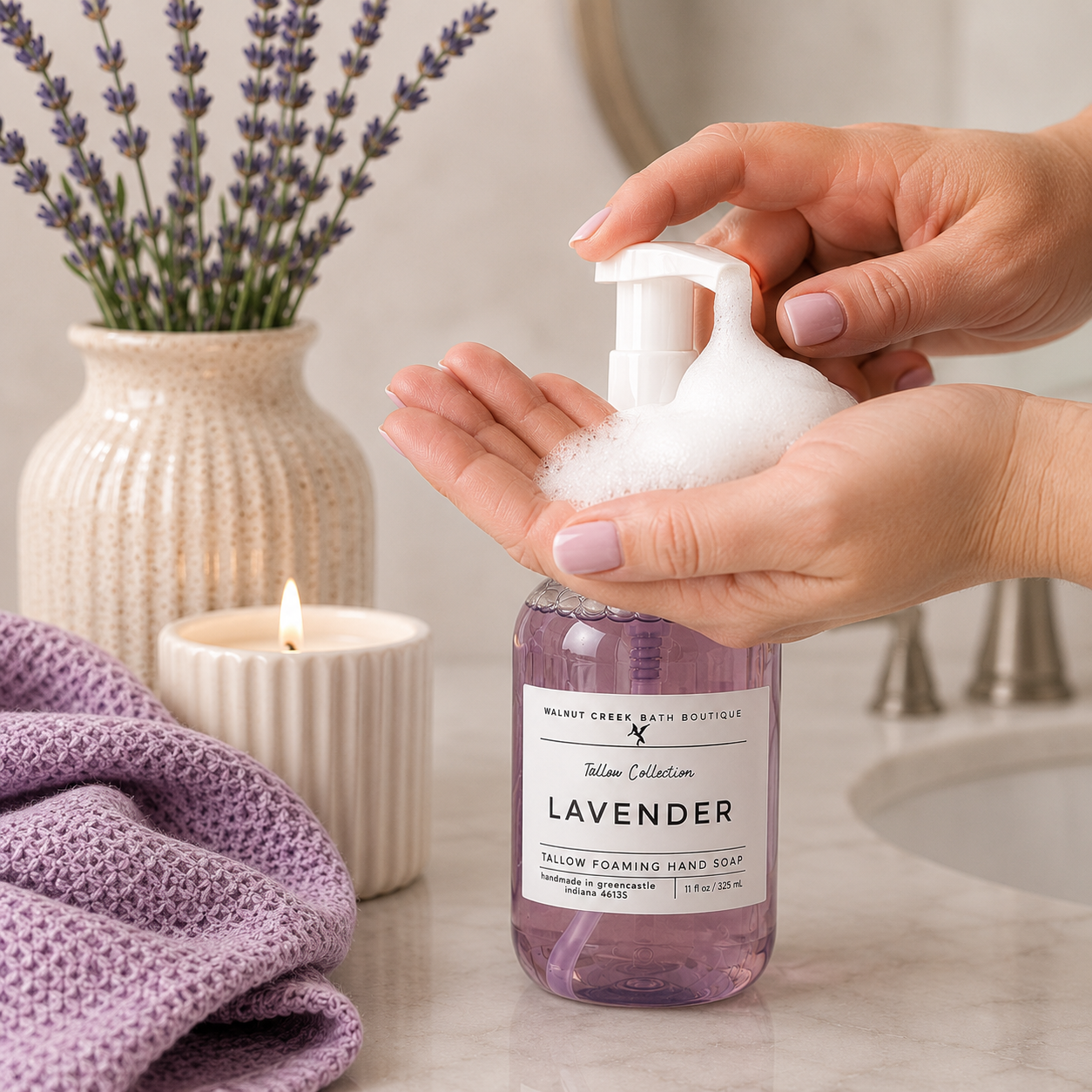 A bottle of lavender salon foaming hand soap being dispensed with a white pump into two hands, with lavender stems in a vase, a lit candle, and a purple towel on a bathroom counter.